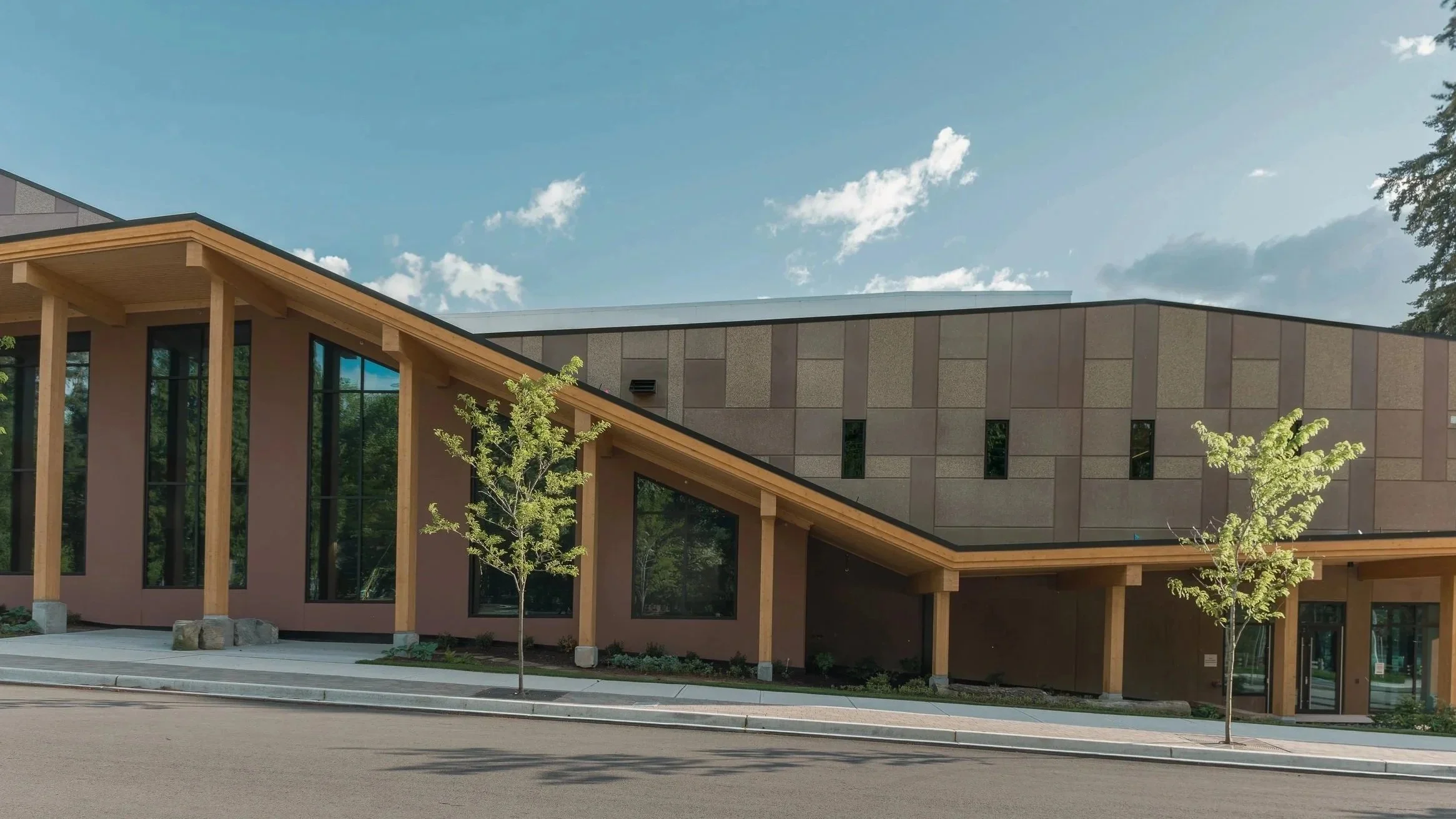 Modern building with sloped roof, large windows, and wooden supports, surrounded by small trees on a sunny day.