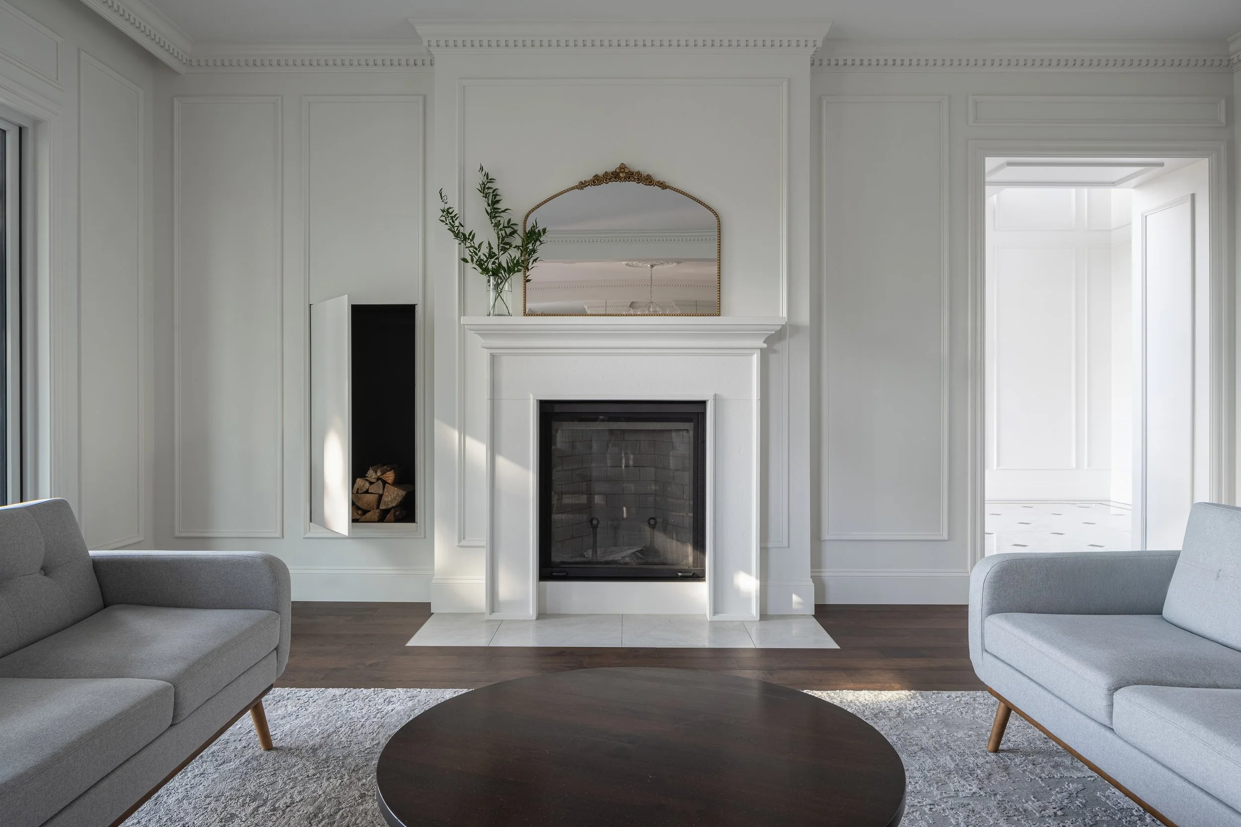 Interior design of a Living room with a fireplace, gray sofas, a dark wooden coffee table, a mirror above the fireplace, a vase with greenery, and a doorway leading to a brightly lit room.