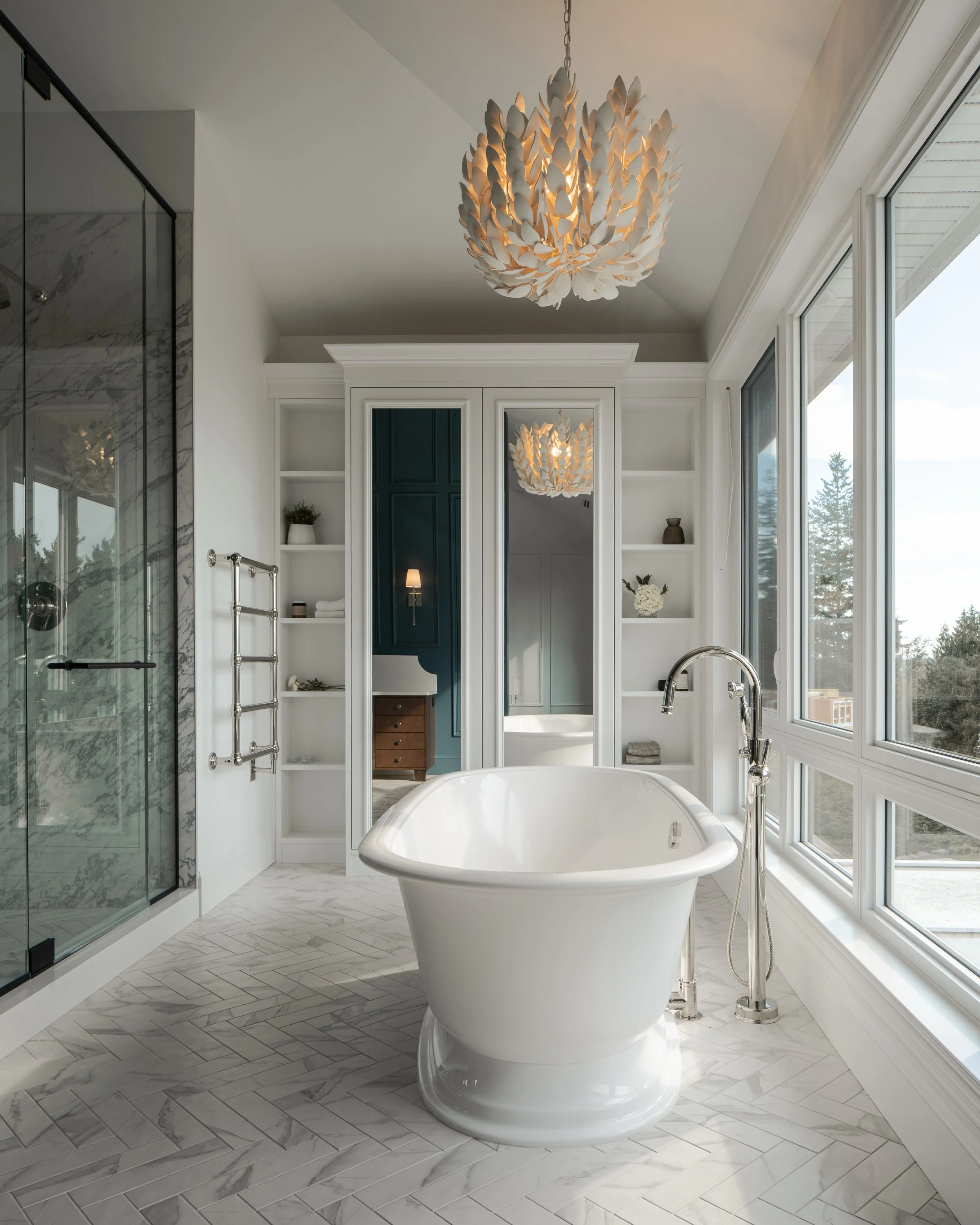 Interior design of a Modern bathroom with a white freestanding bathtub, large windows, marble tile flooring, and a chandelier with leaf-like design.