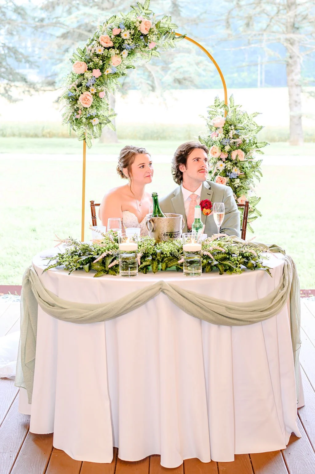 A newlywed couple sits at a decorated wedding table with greenery, candles, and drinks, in front of a floral arch outdoors.