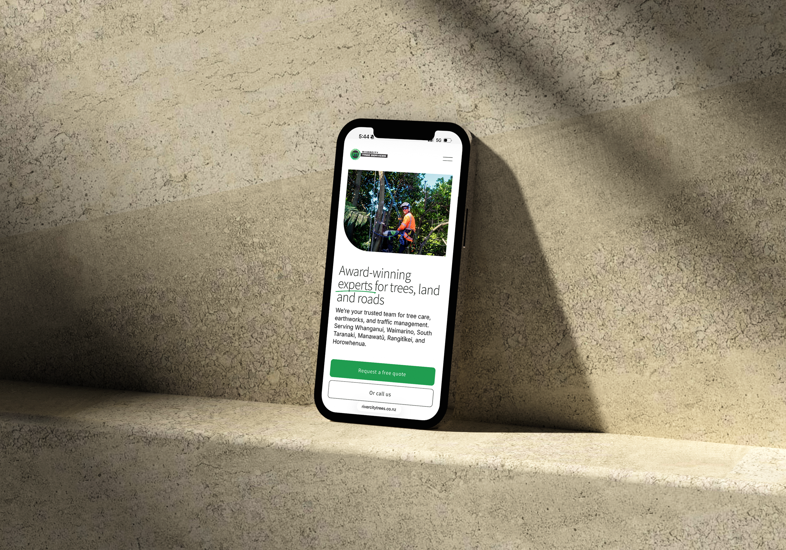 A smartphone displaying a website for a tree care company. The website features a photo of a worker in safety gear and a helmet, working among trees, with text highlighting their expertise in trees, land, and roads.