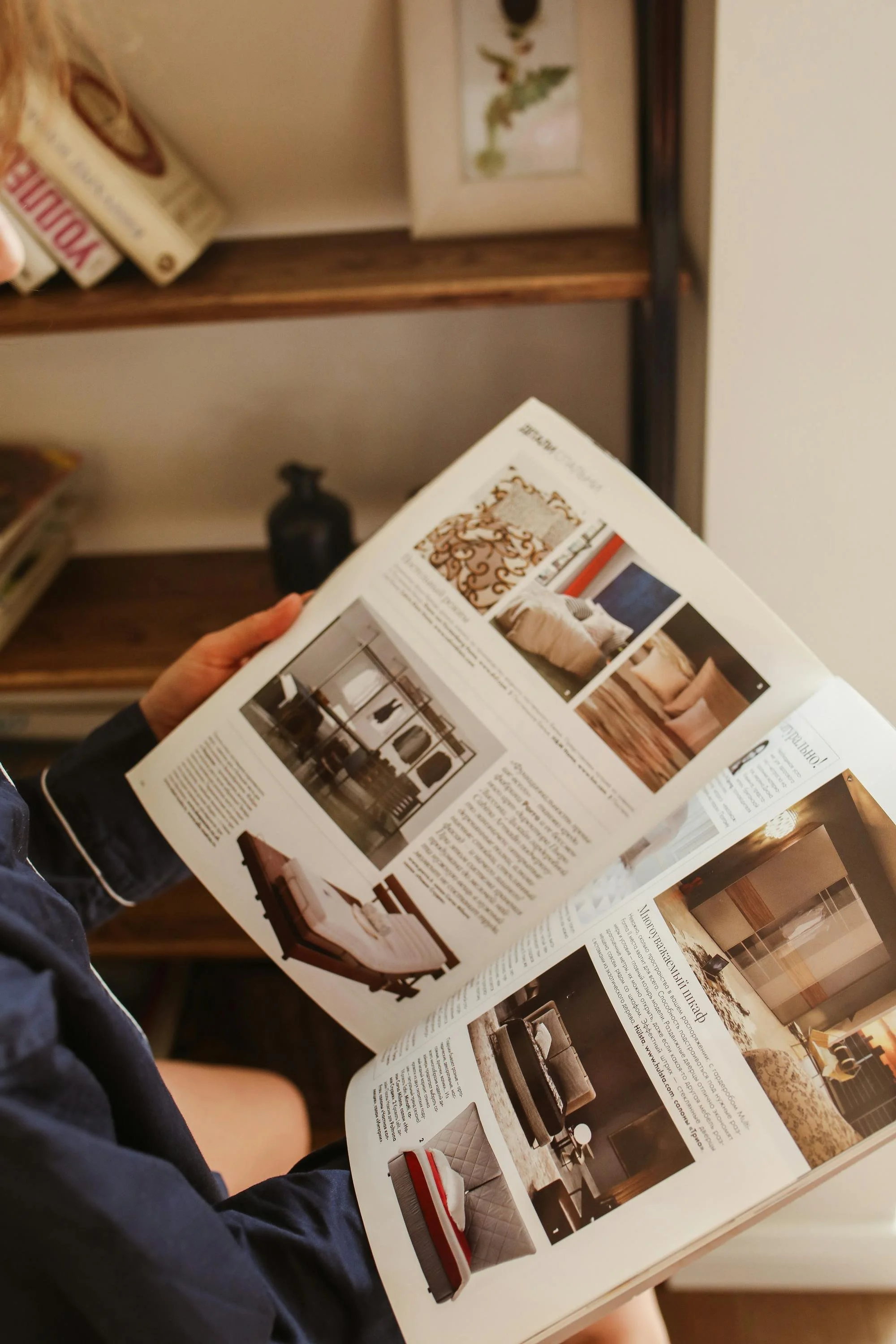 Person reading a magazine featuring furniture and interior design images, sitting near a wooden bookshelf.