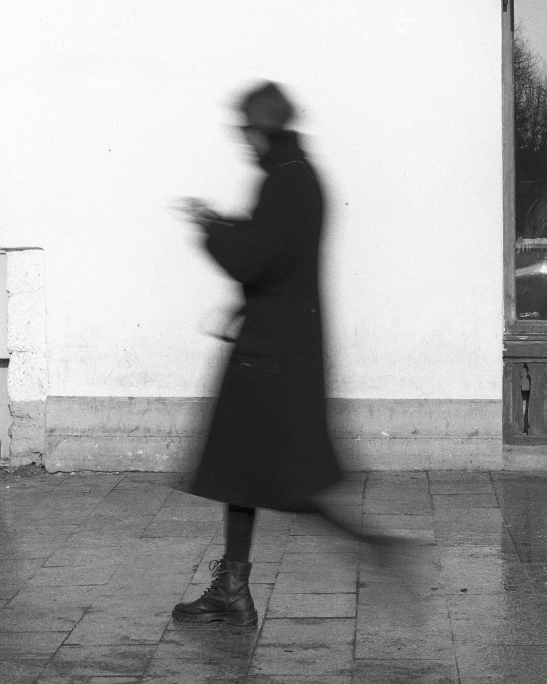 A person walking by a wall, blurred motion, wearing boots, a long coat, and a hat, in black and white.