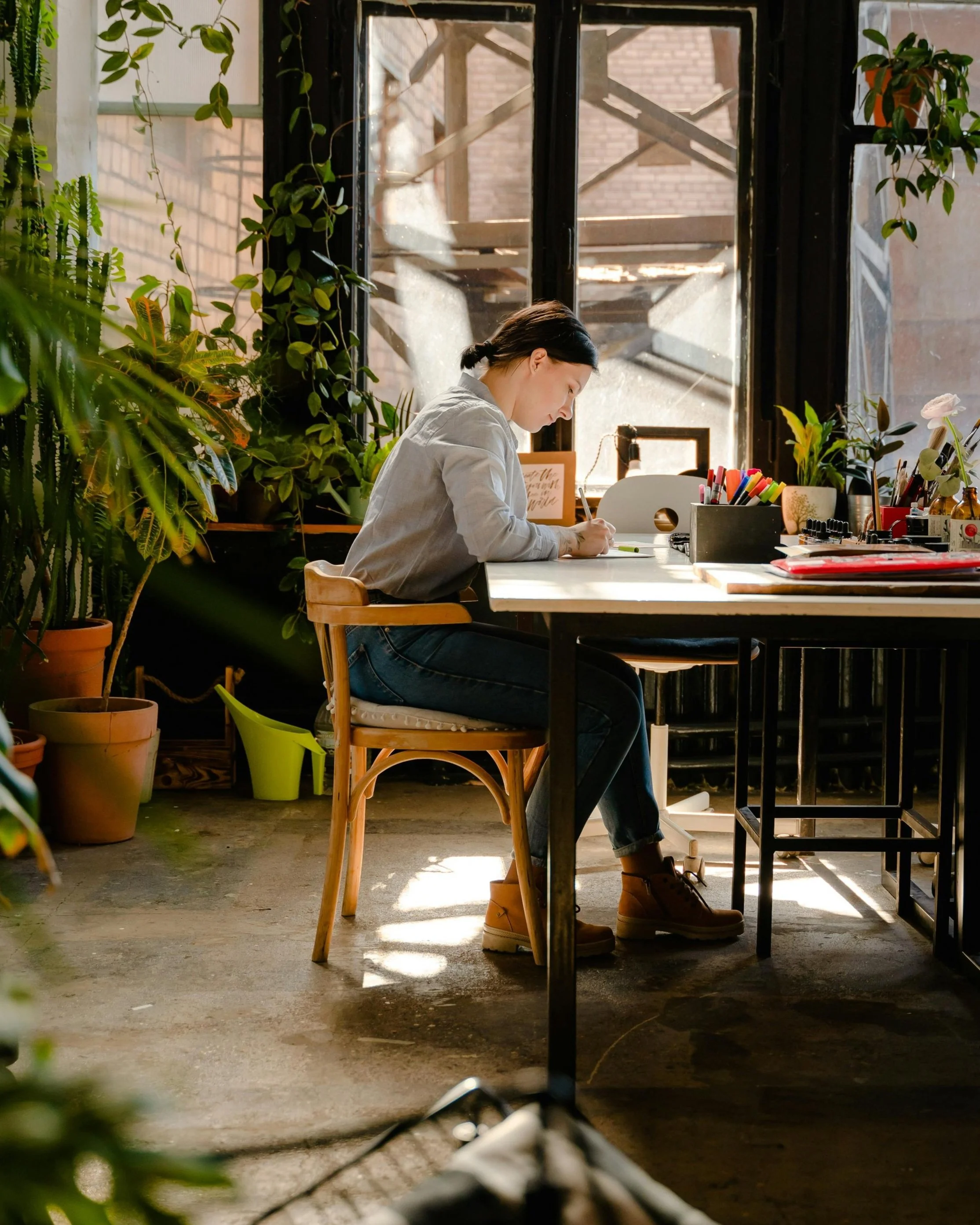 A woman sitting at a desk in a well-lit room surrounded by plants, working on a notebook.