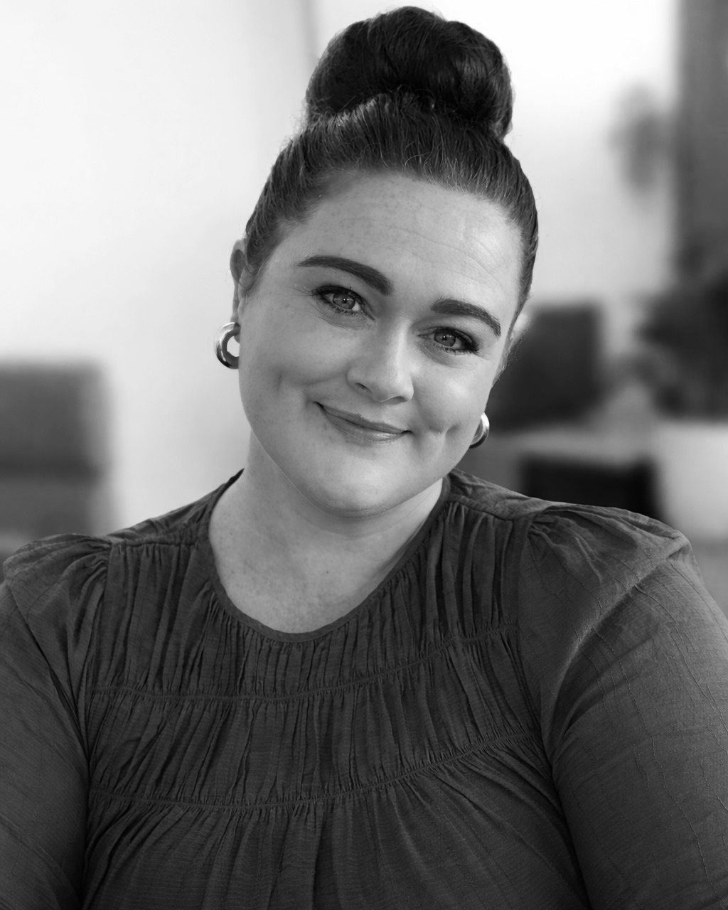 Black-and-white portrait of a woman with dark hair in a high bun, smiling, wearing earrings and a textured blouse, in an indoor setting.
