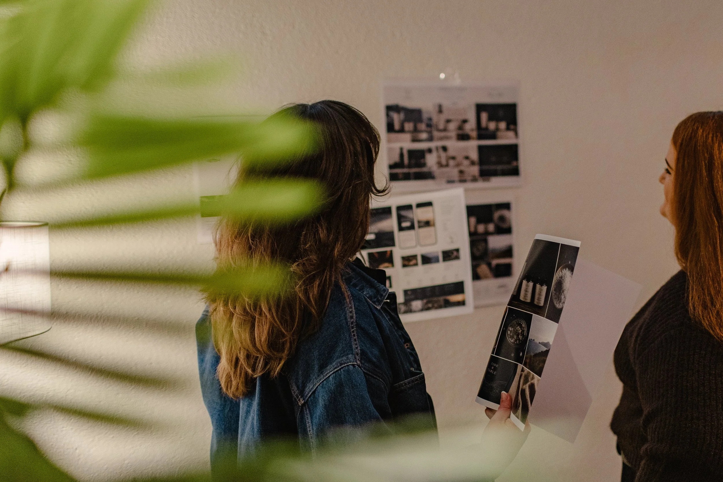 Two women are standing and discussing in an office with visual materials and a wall display behind them, partially obscured by a blurry green plant in the foreground.