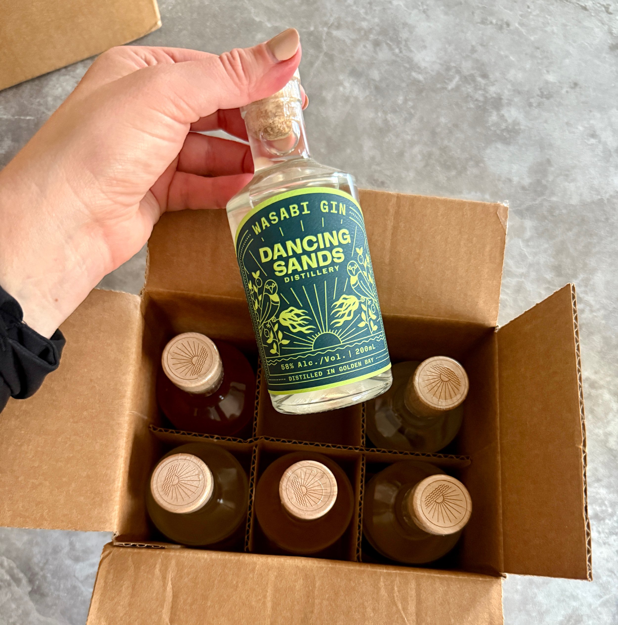 Hand holding a bottle of Wasabi Gin labeled DANCING SANDS, inside a cardboard box containing multiple bottles of similar size and shape with cork stoppers.