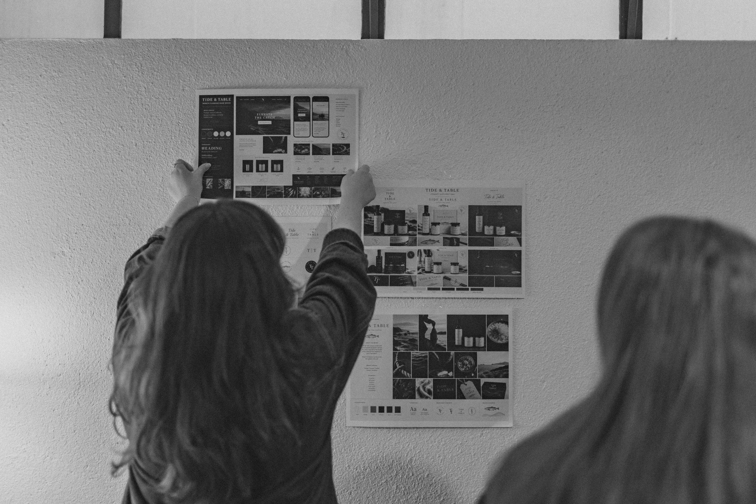 A person with long hair and a jacket pointing at a design poster on a wall, with other people observing the display.