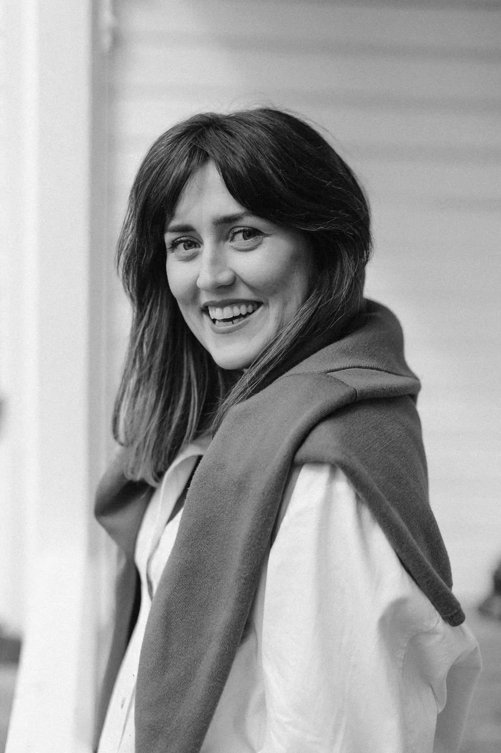 A black-and-white photo of a woman with long hair, smiling, wearing a jacket over a shirt, standing indoors.