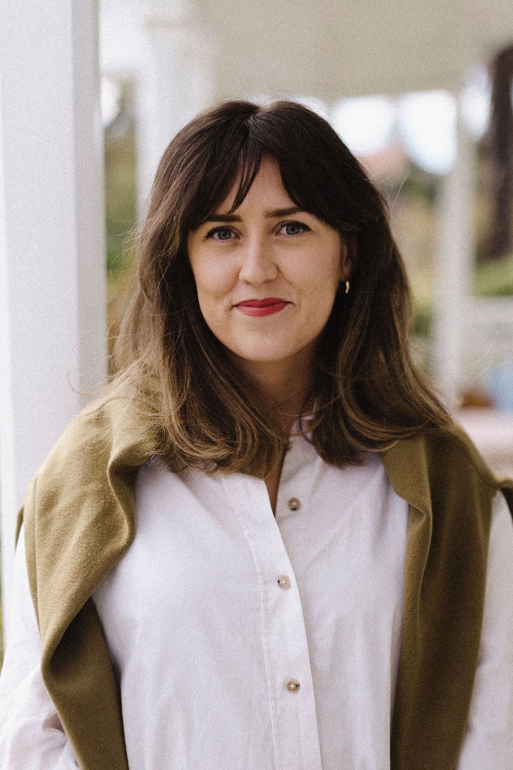 A woman with shoulder-length brown hair, wearing a white blouse with gold buttons and a brown coat draped over her shoulders, standing outside near a white porch railing with a blurred background.