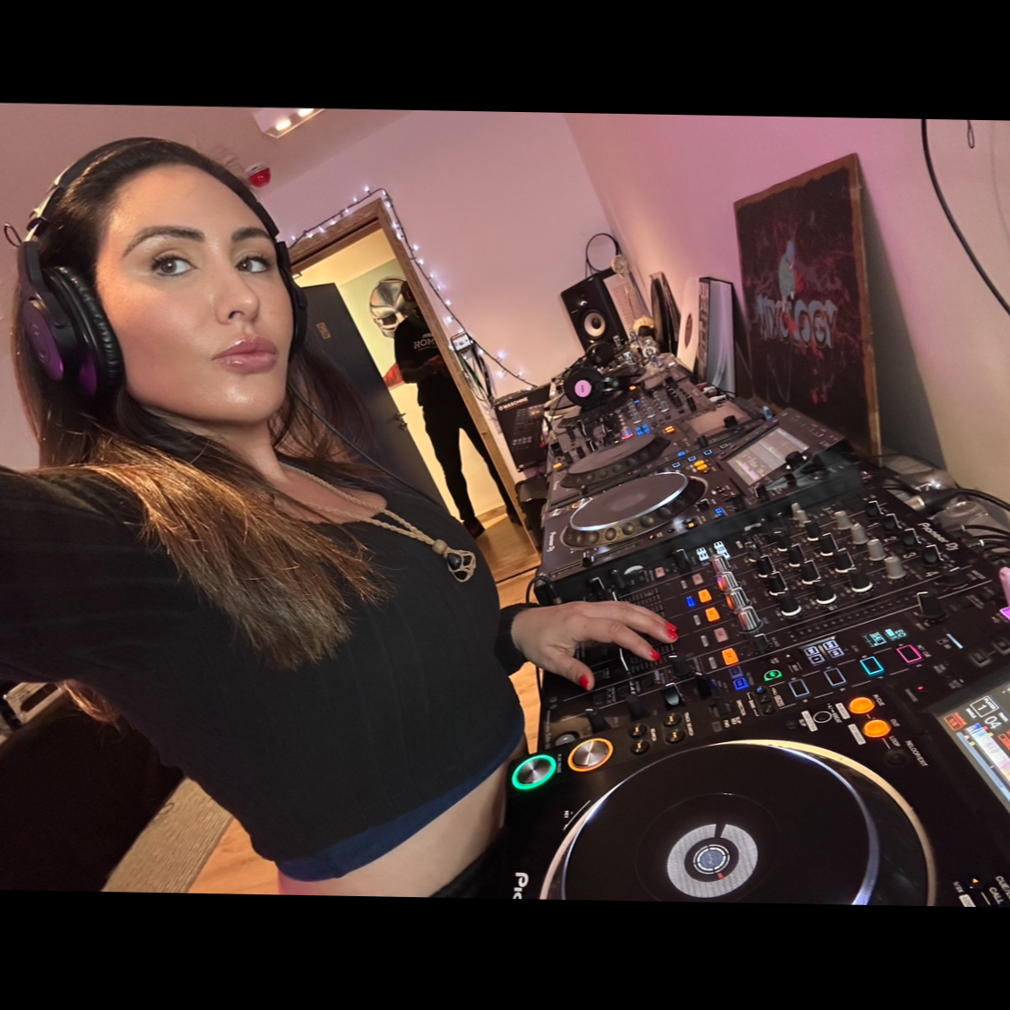 A woman with long brown hair wearing headphones and a black top standing behind DJ equipment in a room with pink walls, string lights, and a side painting.