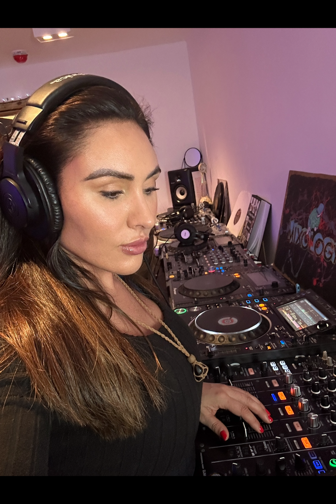 A female DJ wearing headphones, mixing music on a professional DJ controller in a dimly lit room with purple lighting, with various records and speakers in the background.