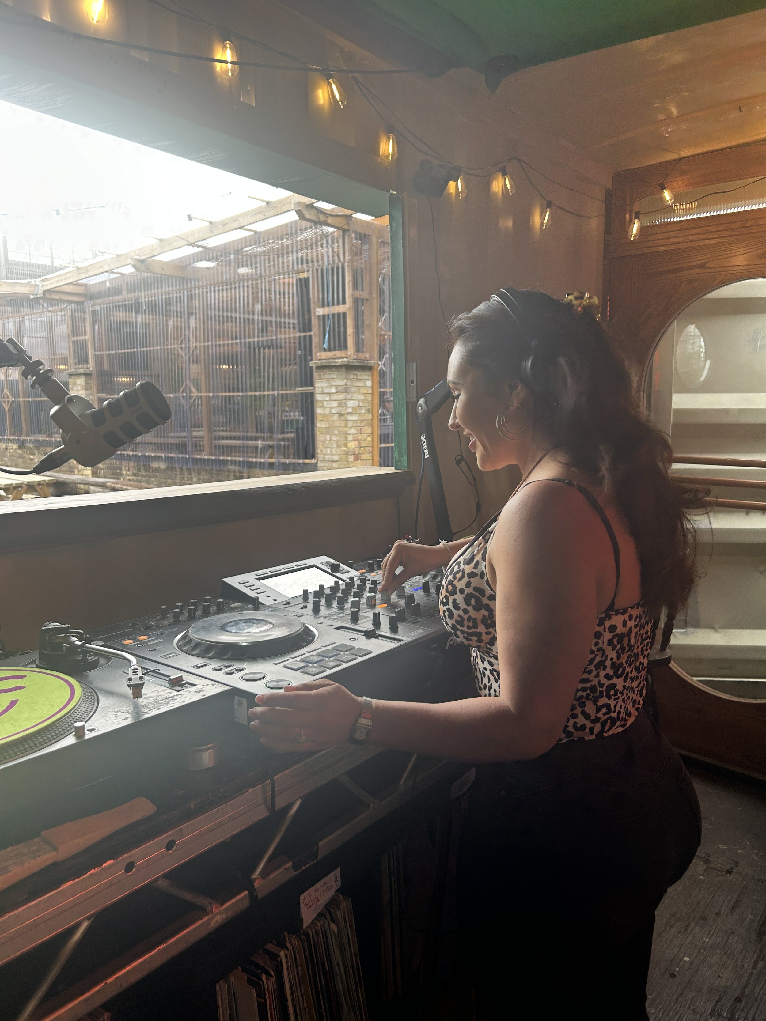 A woman wearing a leopard print top and headphones DJing at a booth with a mixing console, turntable, and microphone, inside a wooden room with string lights.