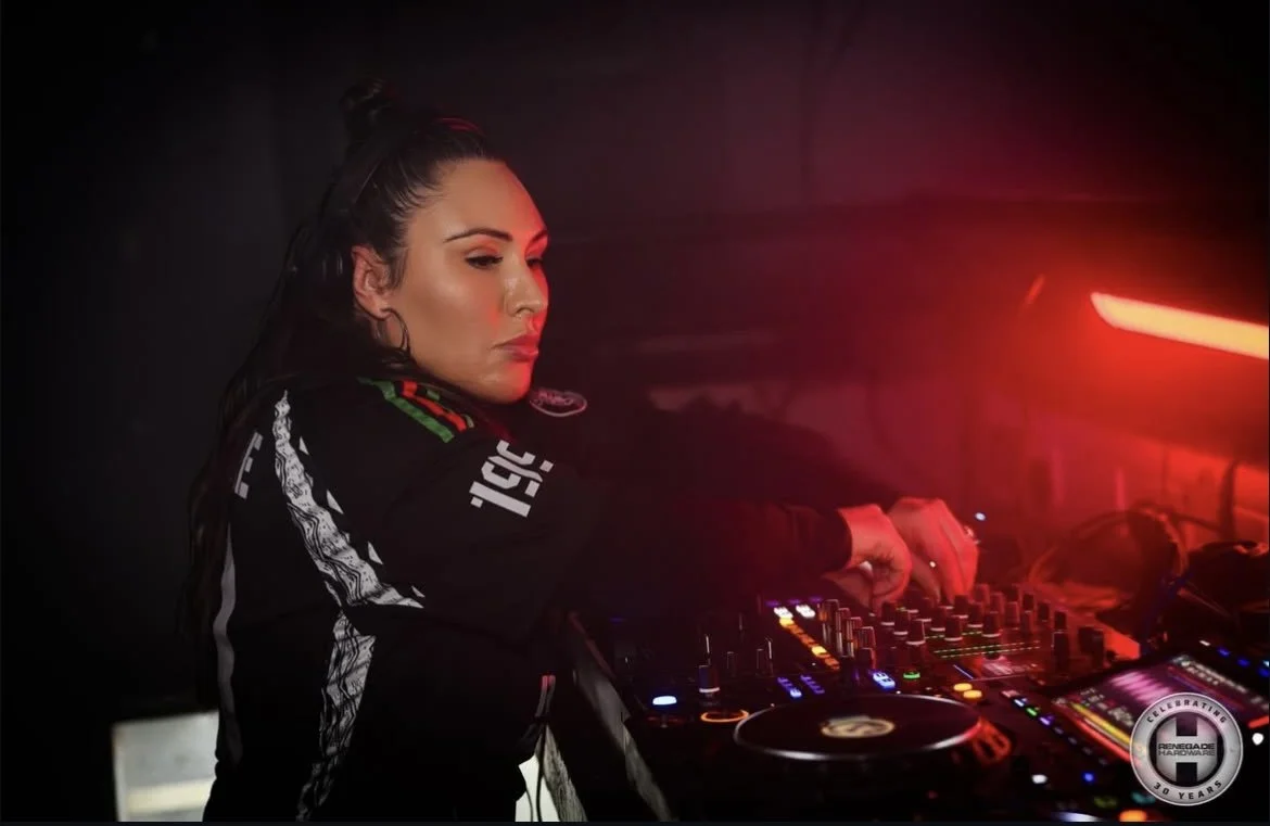 A female DJ with dark hair, wearing a black jacket with racing stripes and logo, stands behind DJ equipment, illuminated by red lighting in a dark setting.