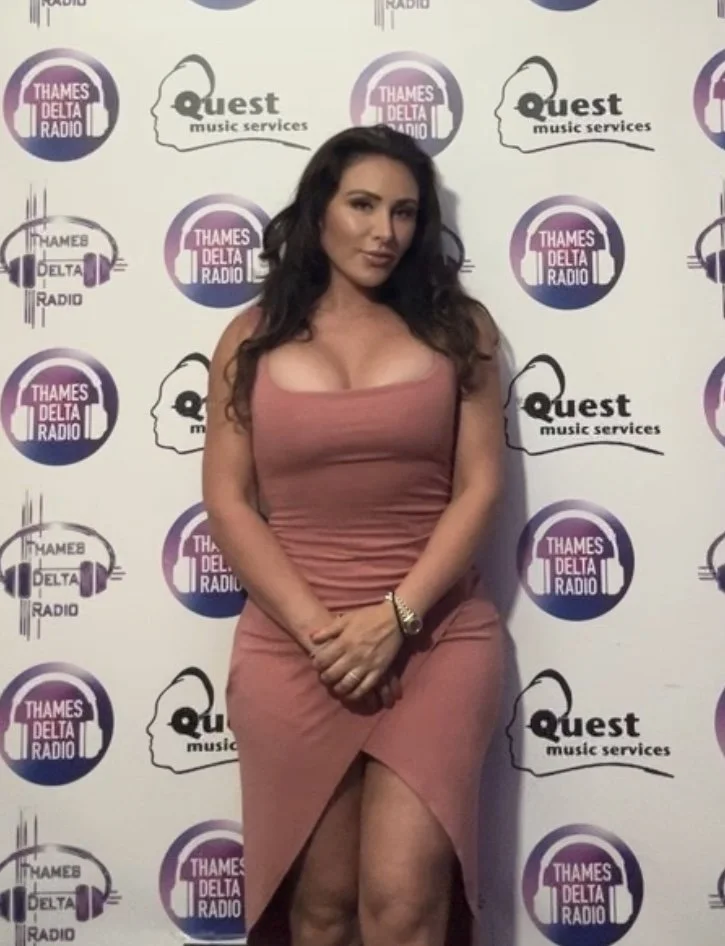 A woman with long dark hair stands in front of a backdrop featuring logos for Thames Delta Radio and Quest Music Services. She is wearing a form-fitting, sleeveless, dusty rose-colored dress and has her hands clasped in front of her.