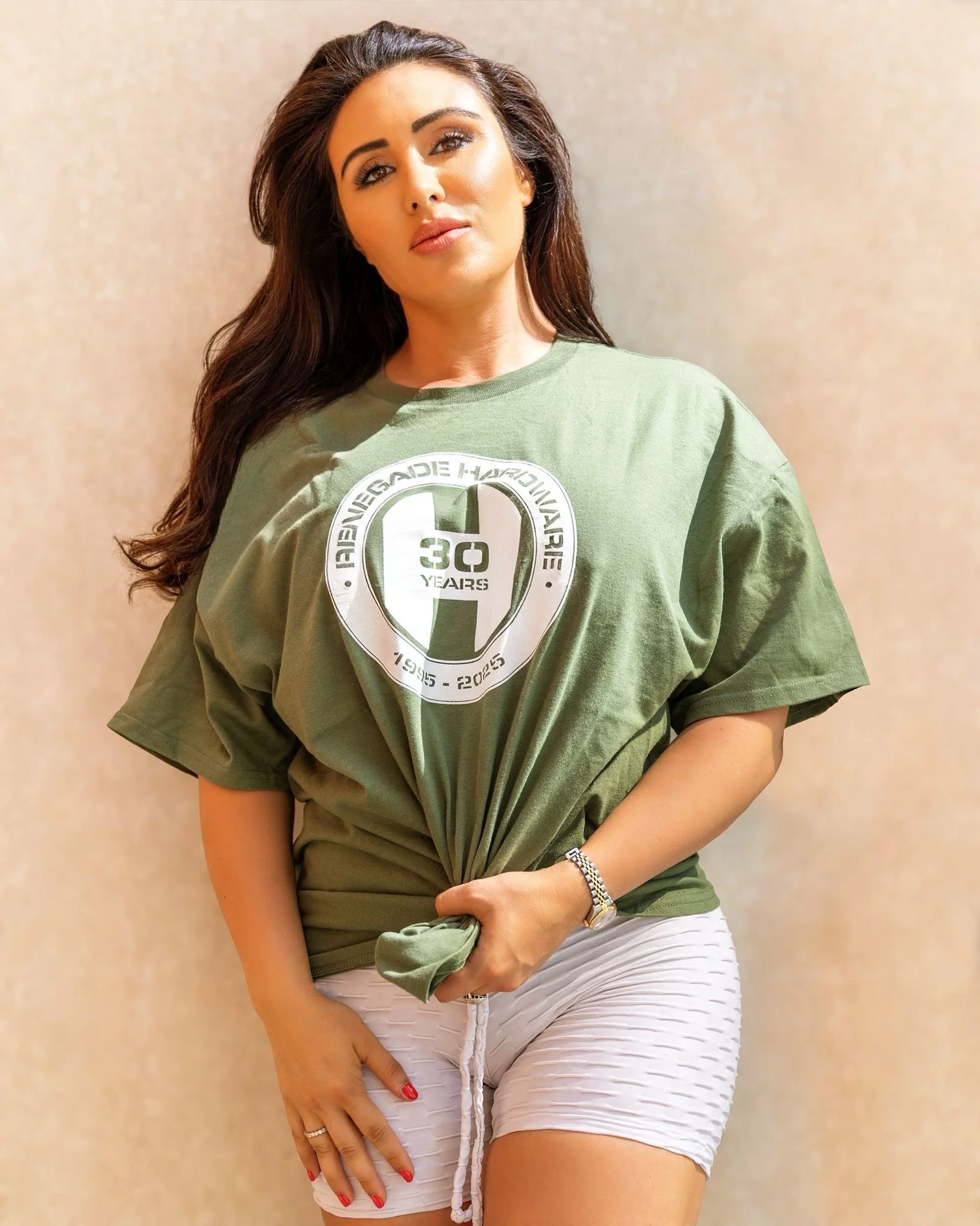 A woman with long dark hair and makeup, wearing a green t-shirt with a logo celebrating 30 years, and white textured shorts, standing against a light-colored wall.
