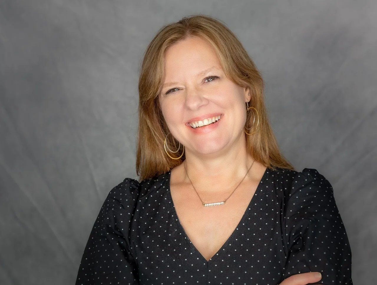 Portrait of a smiling woman with reddish-brown hair, wearing a black polka dot blouse and silver jewelry, against a gray background.