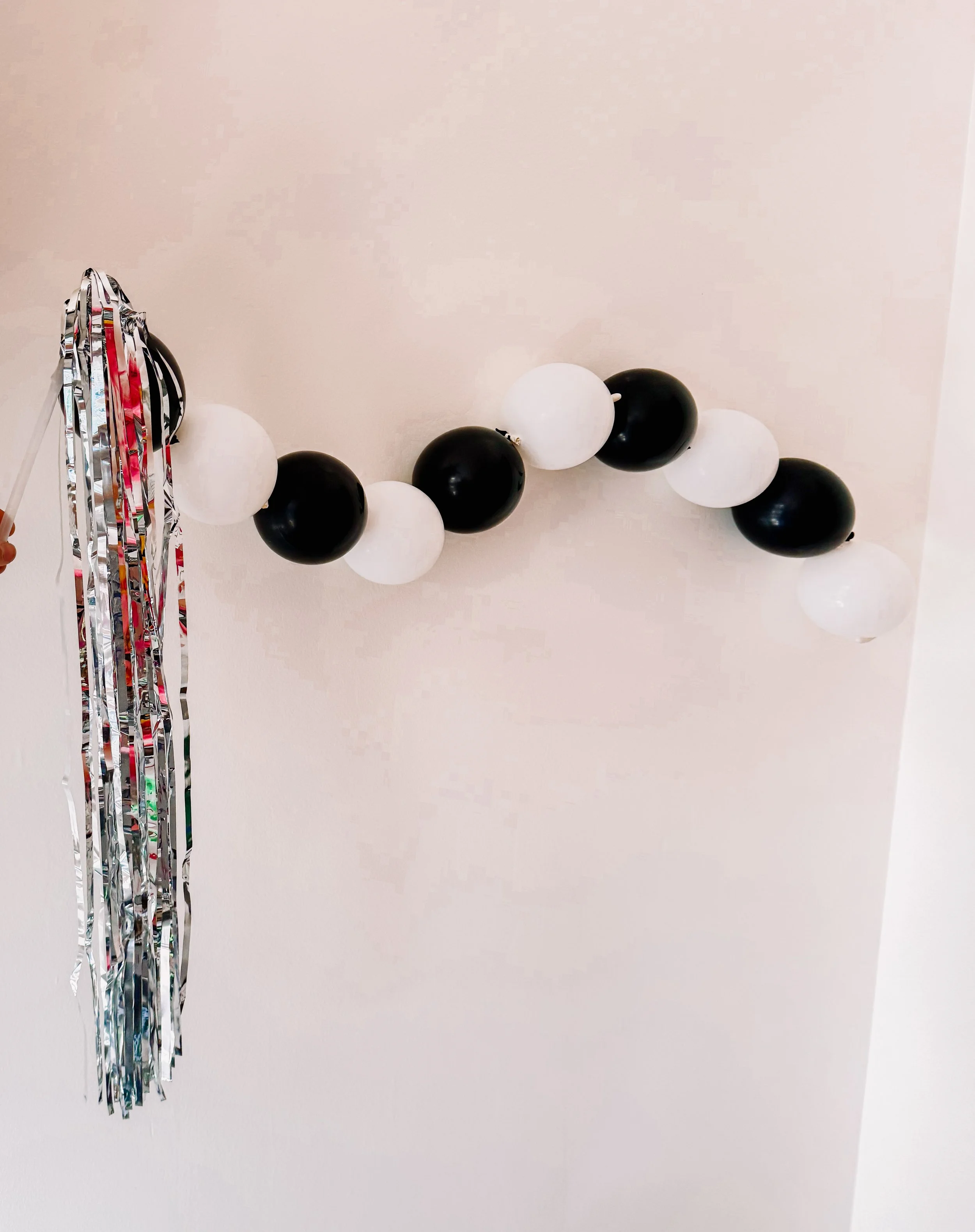 Black and white balloon wand with long metallic silver tinsel fringe, handheld New Year’s party favor for kids and adults.