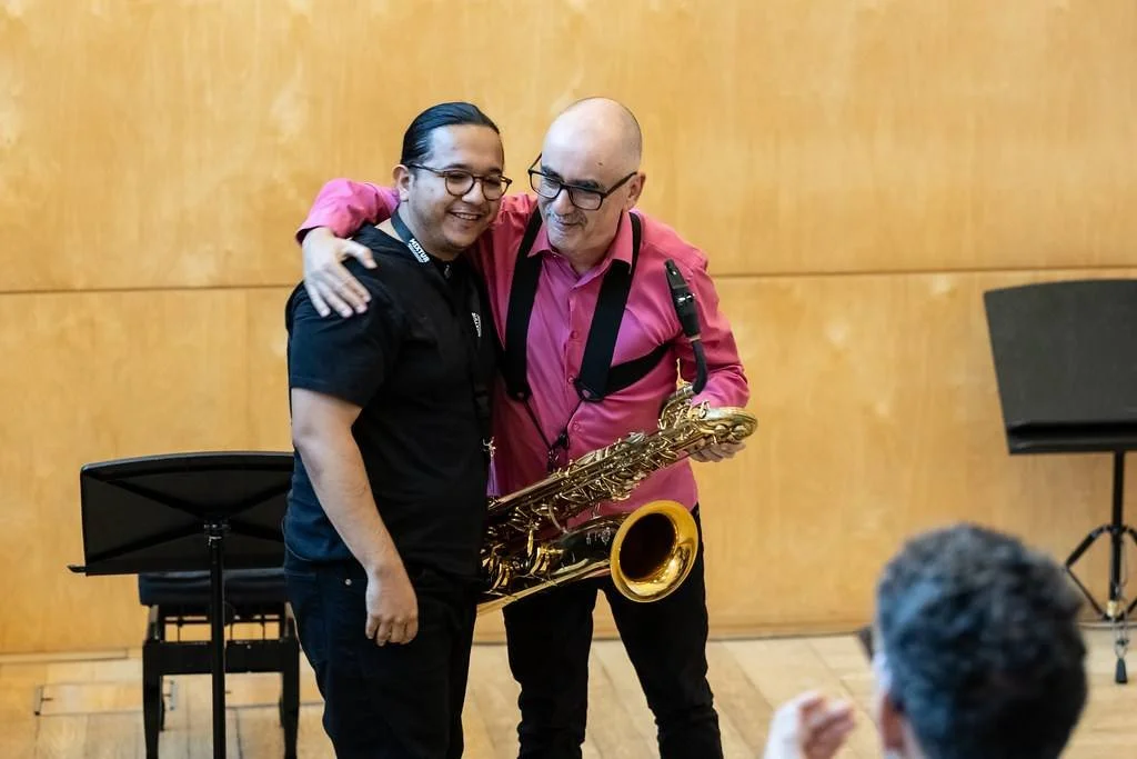 Dos hombres abrazados en un escenario, uno con saxofón, en un ambiente de concierto o espectáculo, con una audiencia visible en primer plano.