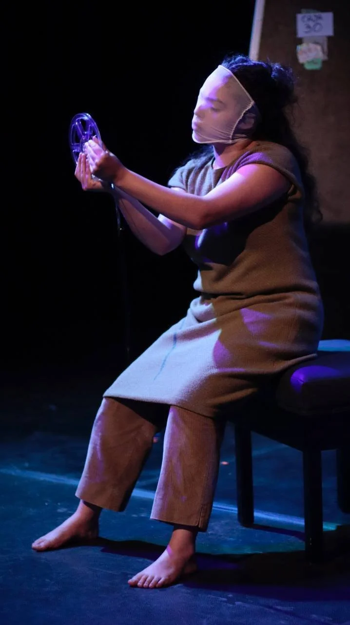 Persona en un escenario, usando una máscara, sosteniendo un objeto circular, sentada en una banca, con fondo oscuro.