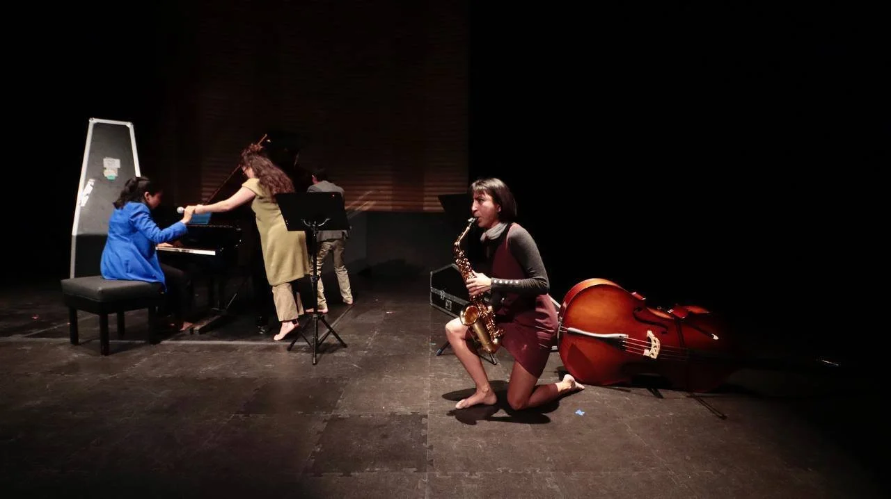 Tres personas en un escenario de teatro tocando instrumentos musicales. Una niña en un piano, una mujer en el saxofón y otra niña entregando algo al pianista. Un contrabajo está acostado en el suelo a la derecha.