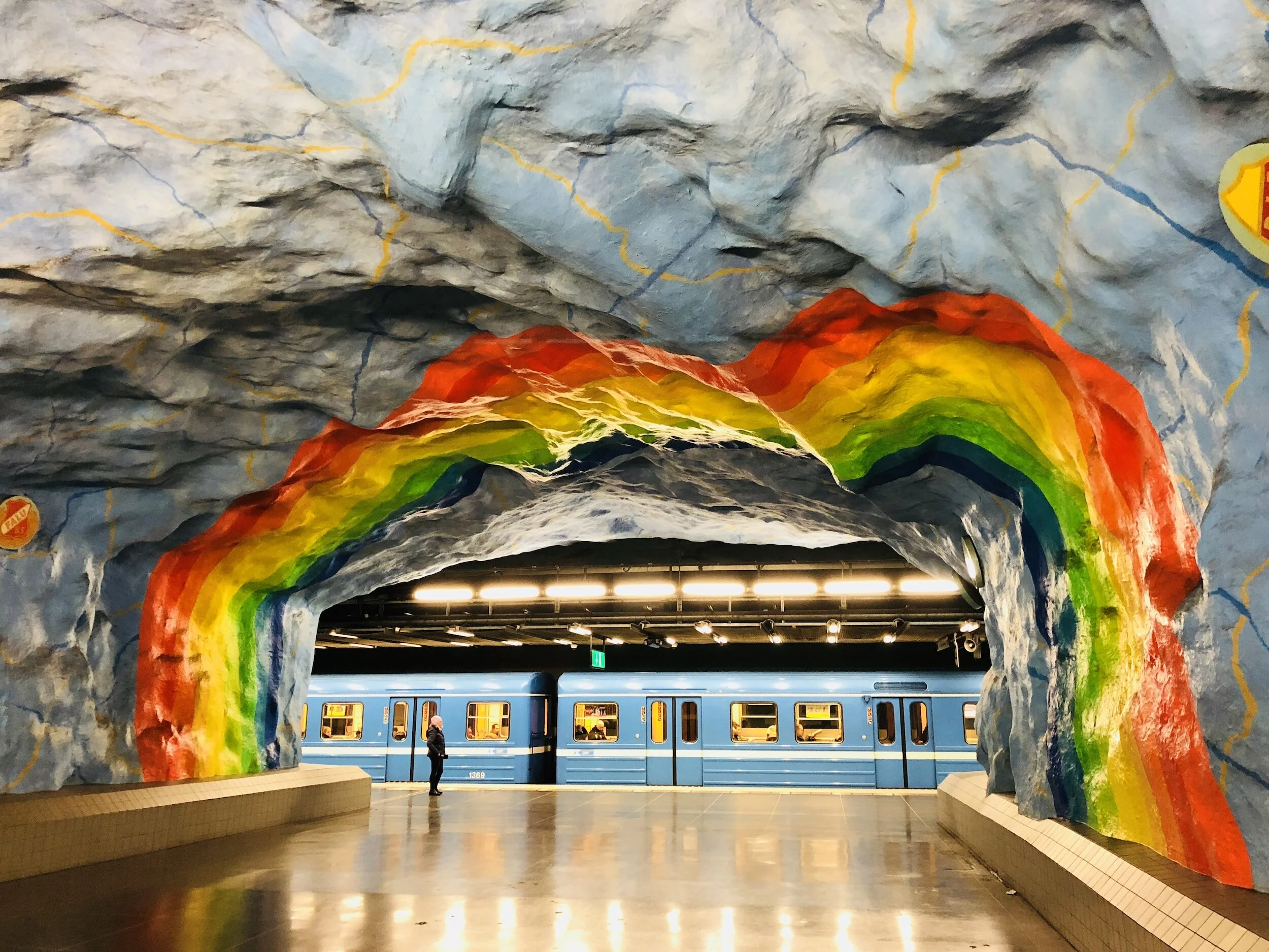 An underground subway station with a vibrant, colorful rock formation painted on the ceiling resembling a rainbow. A blue train is at the platform with passengers visible through the windows, and a person is standing on the platform near the train.