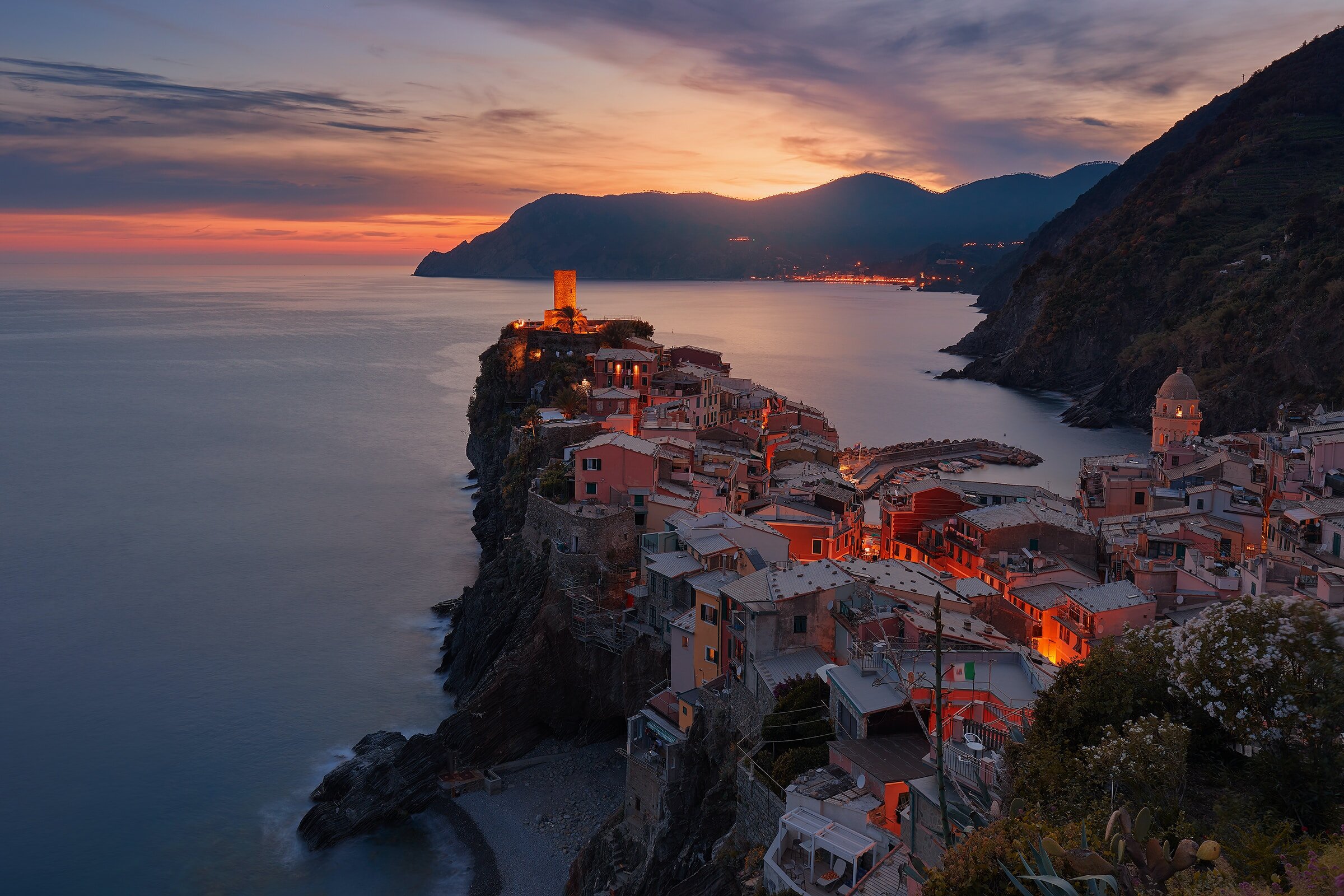 Colorful coastal town at sunset with pink, orange, and purple sky, hillside buildings, and a tower on a rocky promontory by the water.