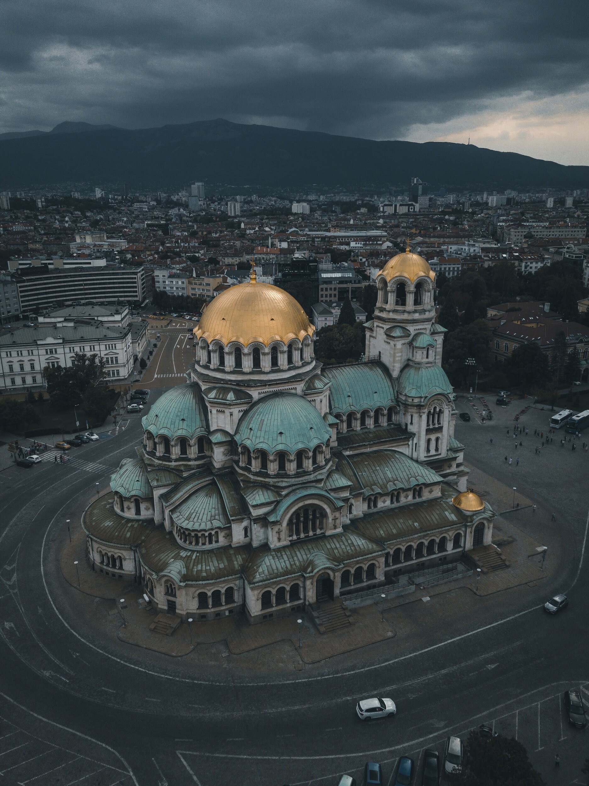 Aerial view of a historic church with green domes and golden accents, set against a cityscape with modern buildings and a mountain range in the background, under a dark cloudy sky.