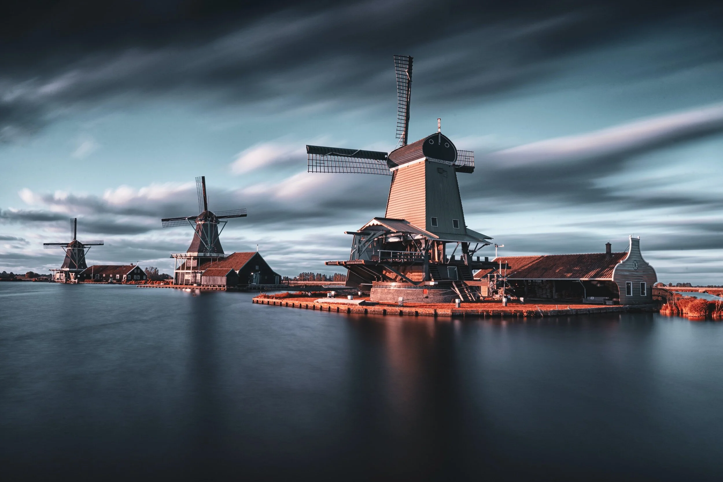 Two traditional Dutch windmills beside a waterway under a cloudy sky during sunset.