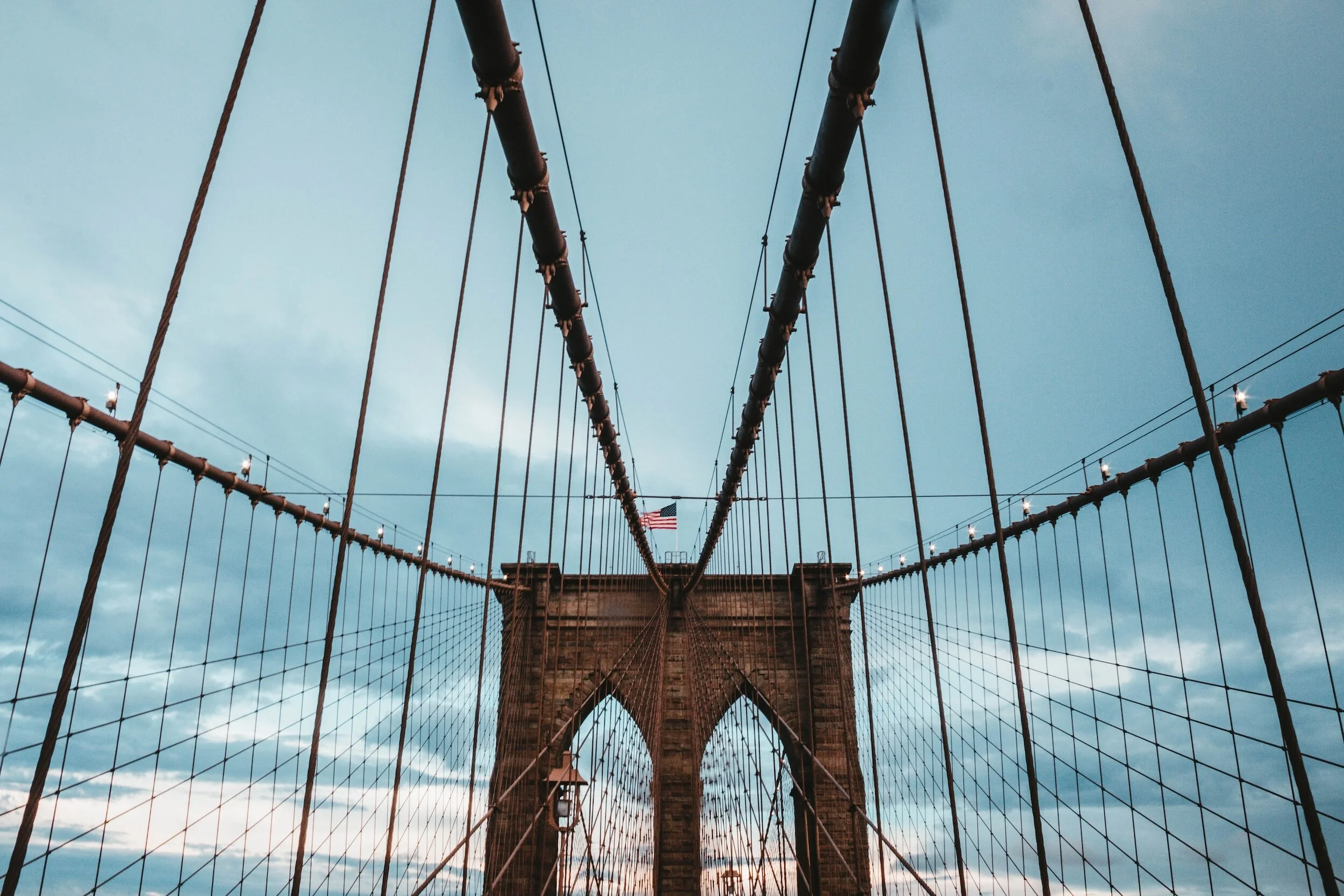 Looking up at the Brooklyn Bridge's stone tower with cables extending outward, against a sky with scattered clouds and an American flag at the top.