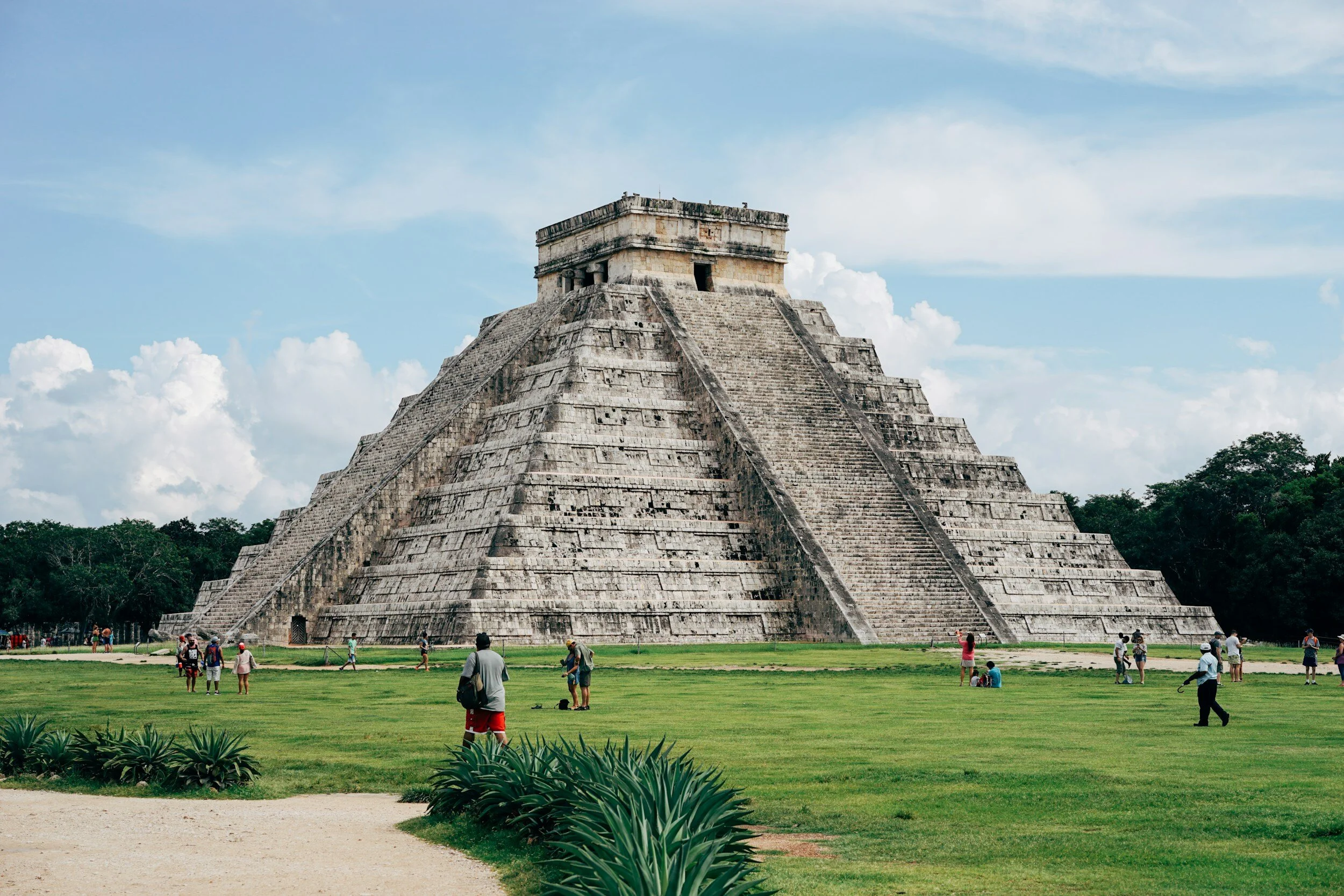 A large stepped pyramid, part of the ancient Mayan city of Chichen Itza, with tourists walking and taking photos in the foreground, under a partly cloudy sky.