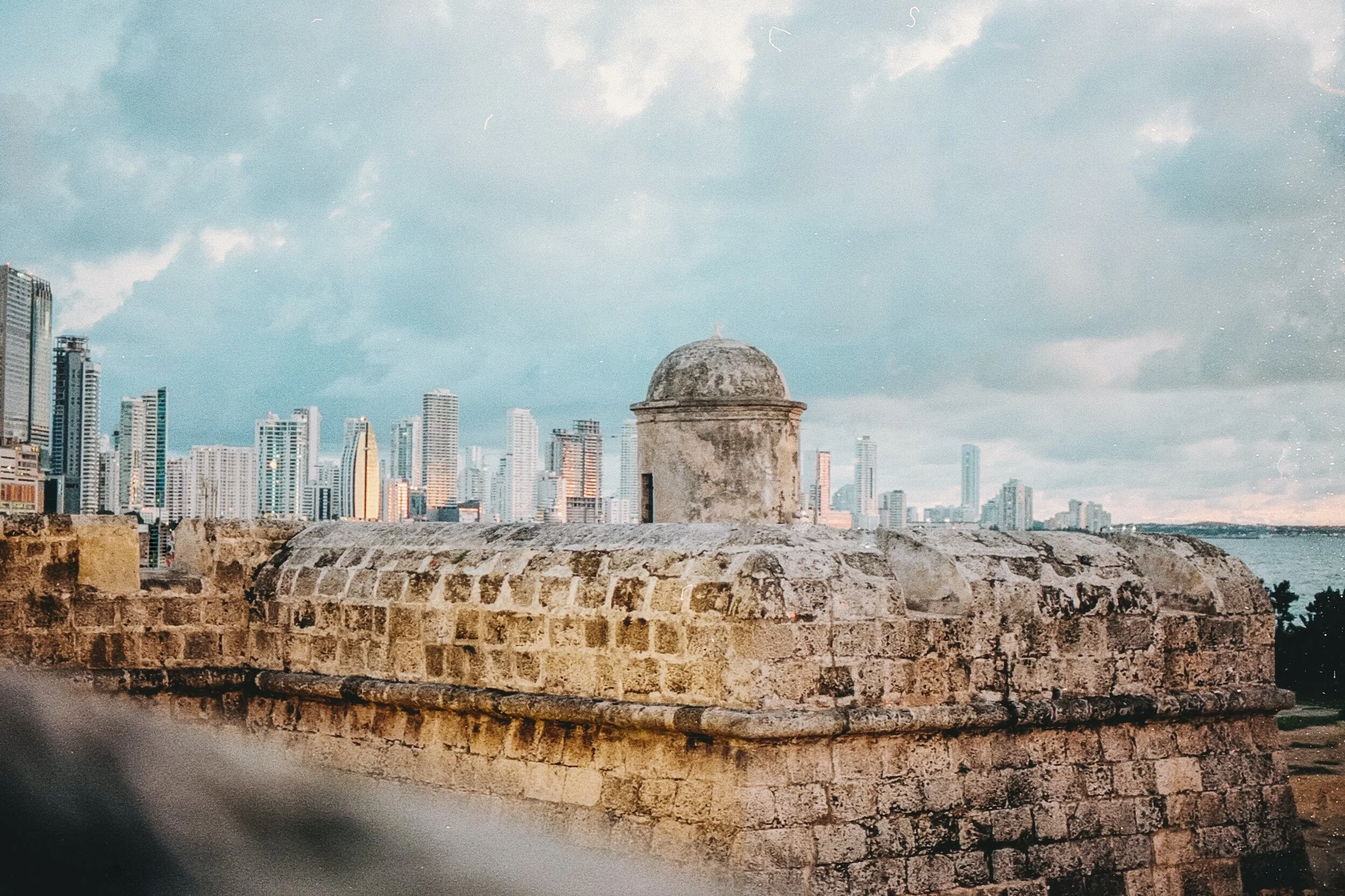 City skyline with historic stone wall and fortress in the foreground, modern skyscrapers in the background, under a partly cloudy sky.