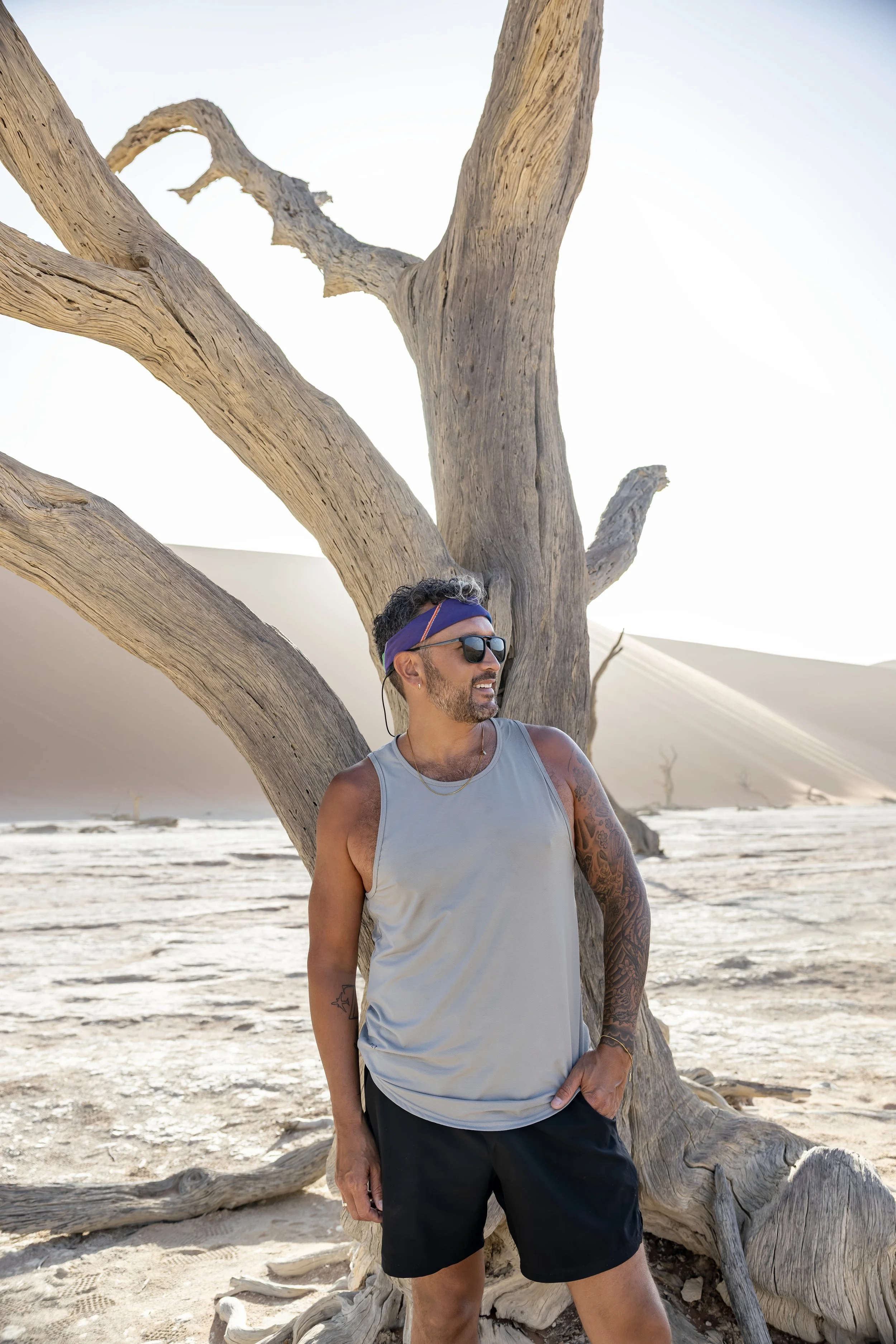 Tommy wearing a sleeveless athletic shirt, shorts, sunglasses, and a headband standing outdoors near a large, twisted dead tree in a sandy desert landscape.
