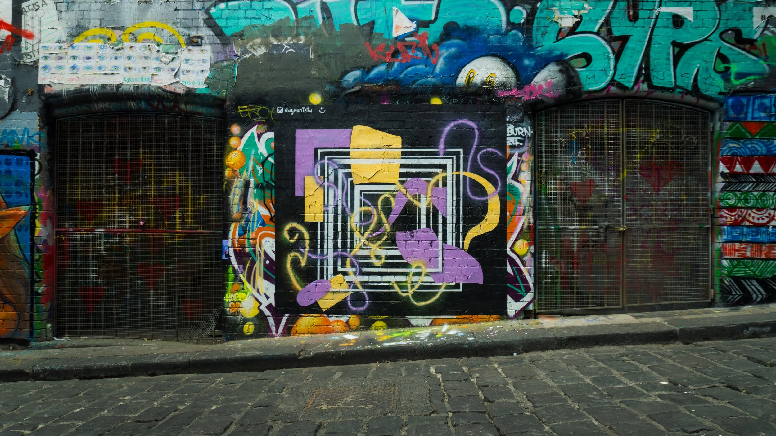 Vibrant street art on a brick wall featuring colorful geometric shapes, swirls, and graffiti with two metal gates on either side.