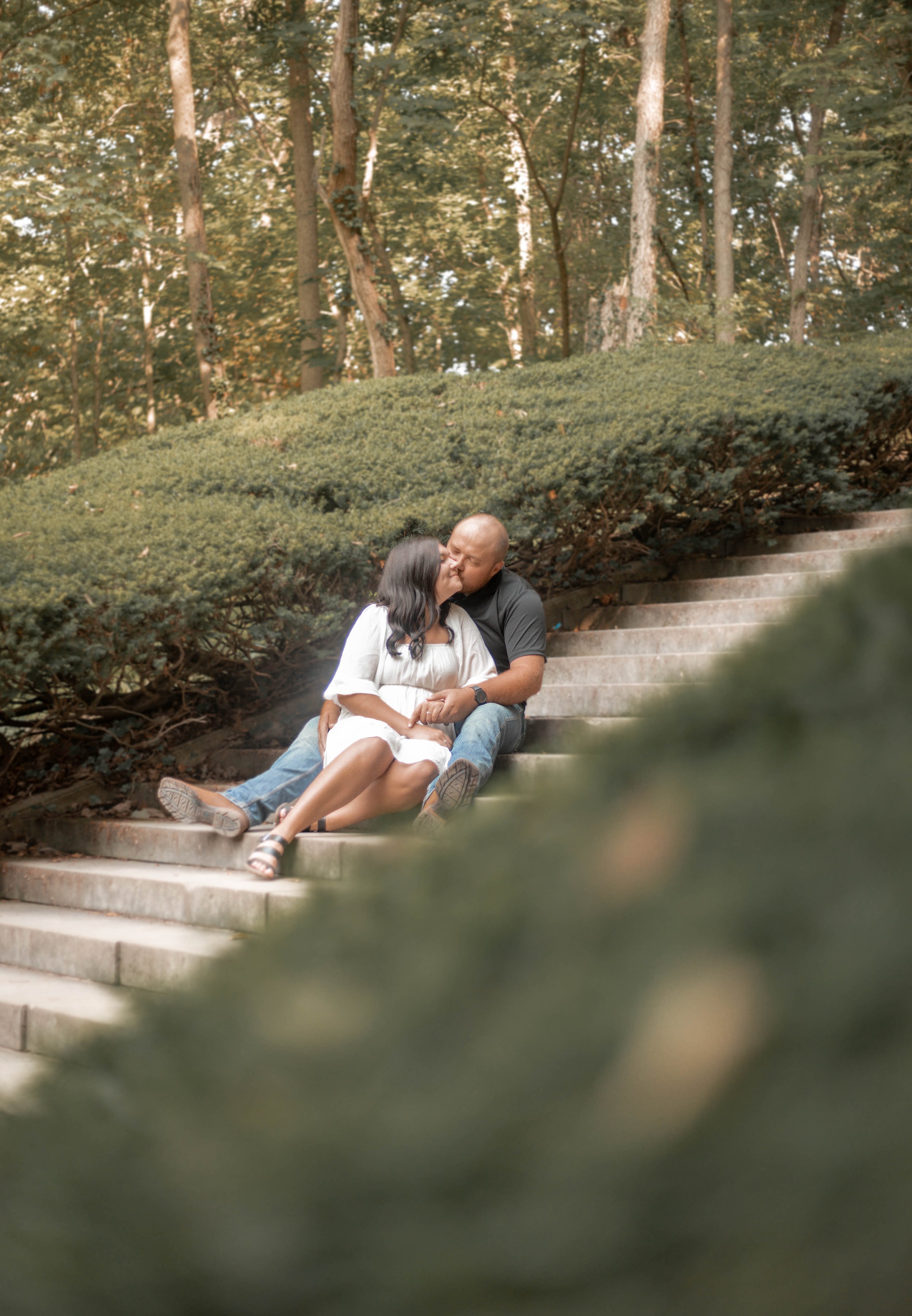 Couple sitting on outdoor stairs sharing a kiss in a wooded park.