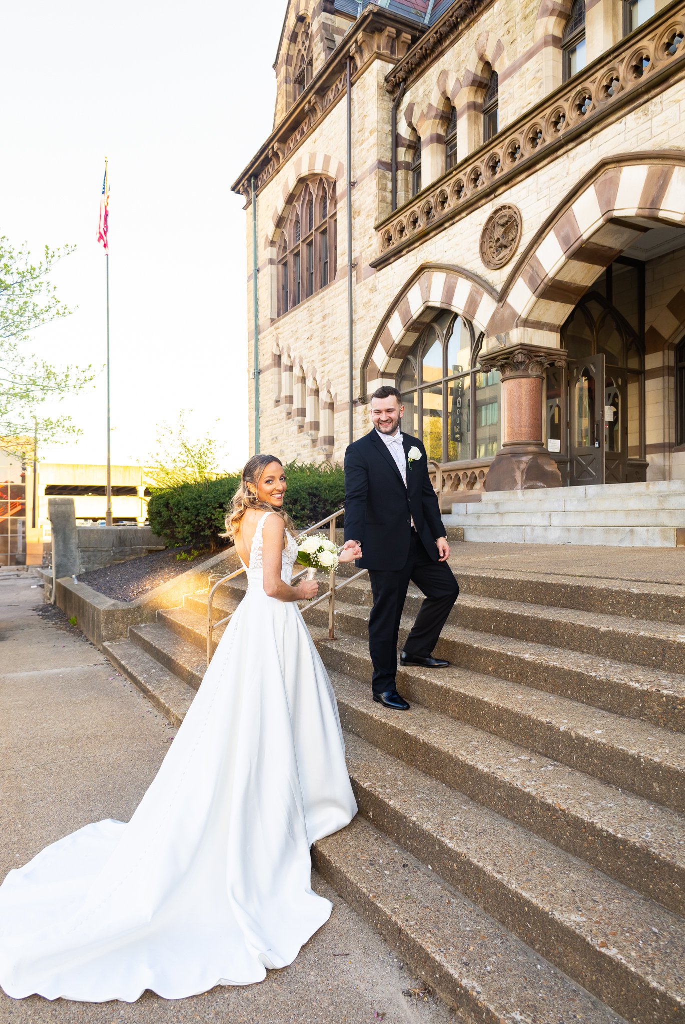A bride and groom standing on stone steps outside a historic building, smiling for a wedding photo. The bride is in a white wedding gown holding a bouquet, and the groom is in a black suit with a white boutonniere.
