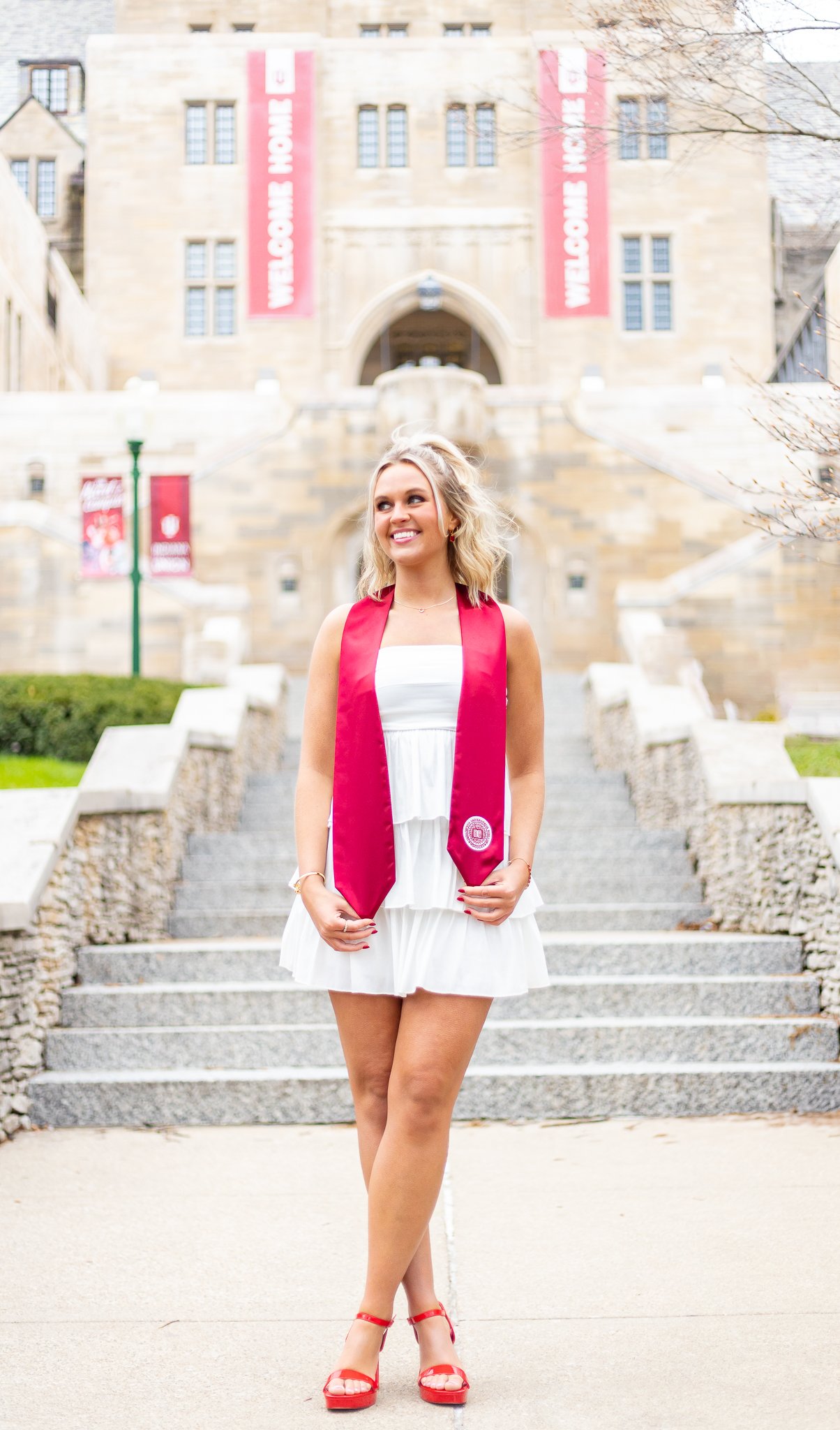 A young woman in a white dress and red heels standing on stone steps in front of a large, historic university building with banners that say 'Welcome Home'.