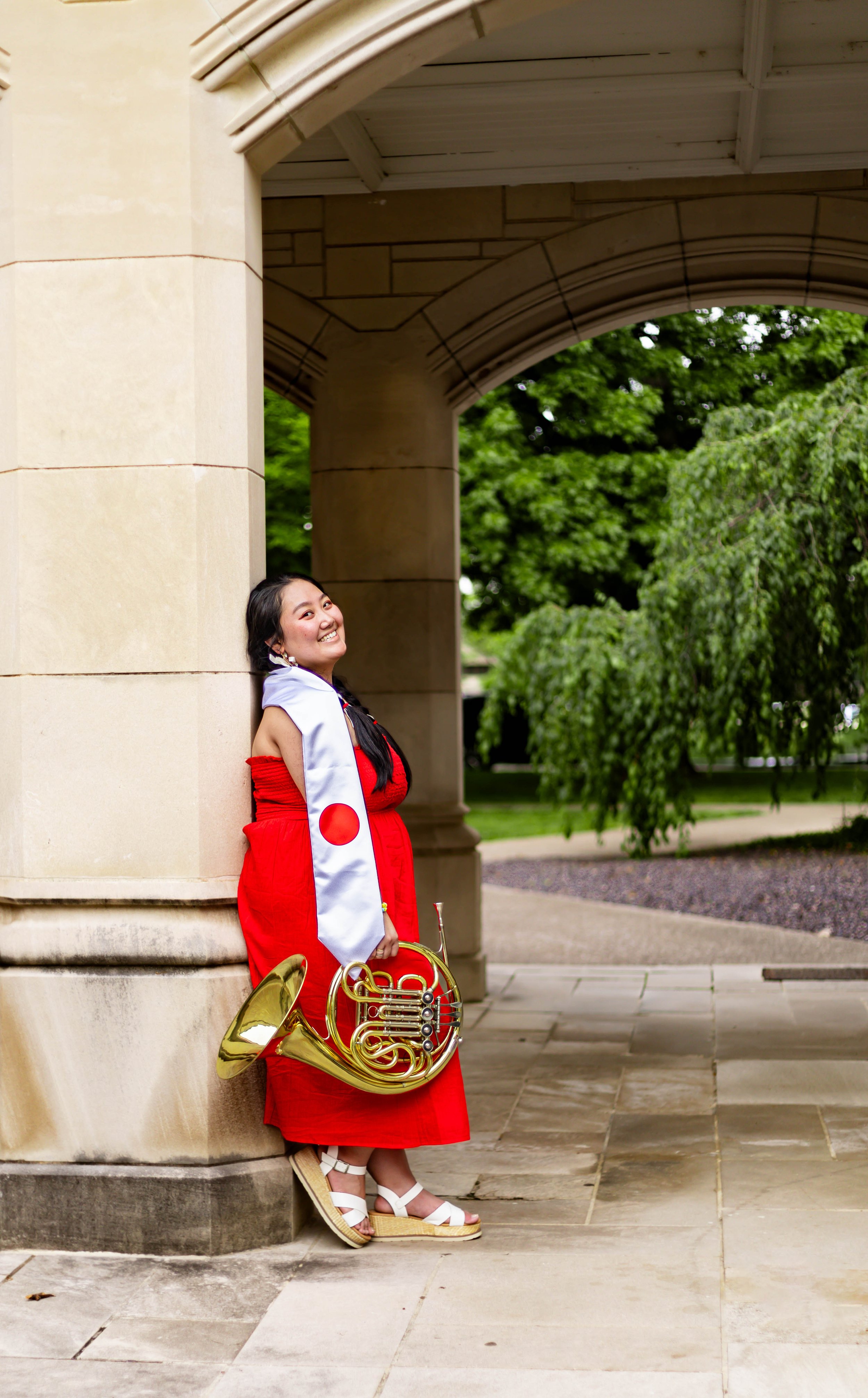 Young woman in a red dress, white scarf with red circle emblem, and white platform sandals, leaning against a stone column under a stone arch, holding a brass French horn, smiling outdoors with green trees in the background.