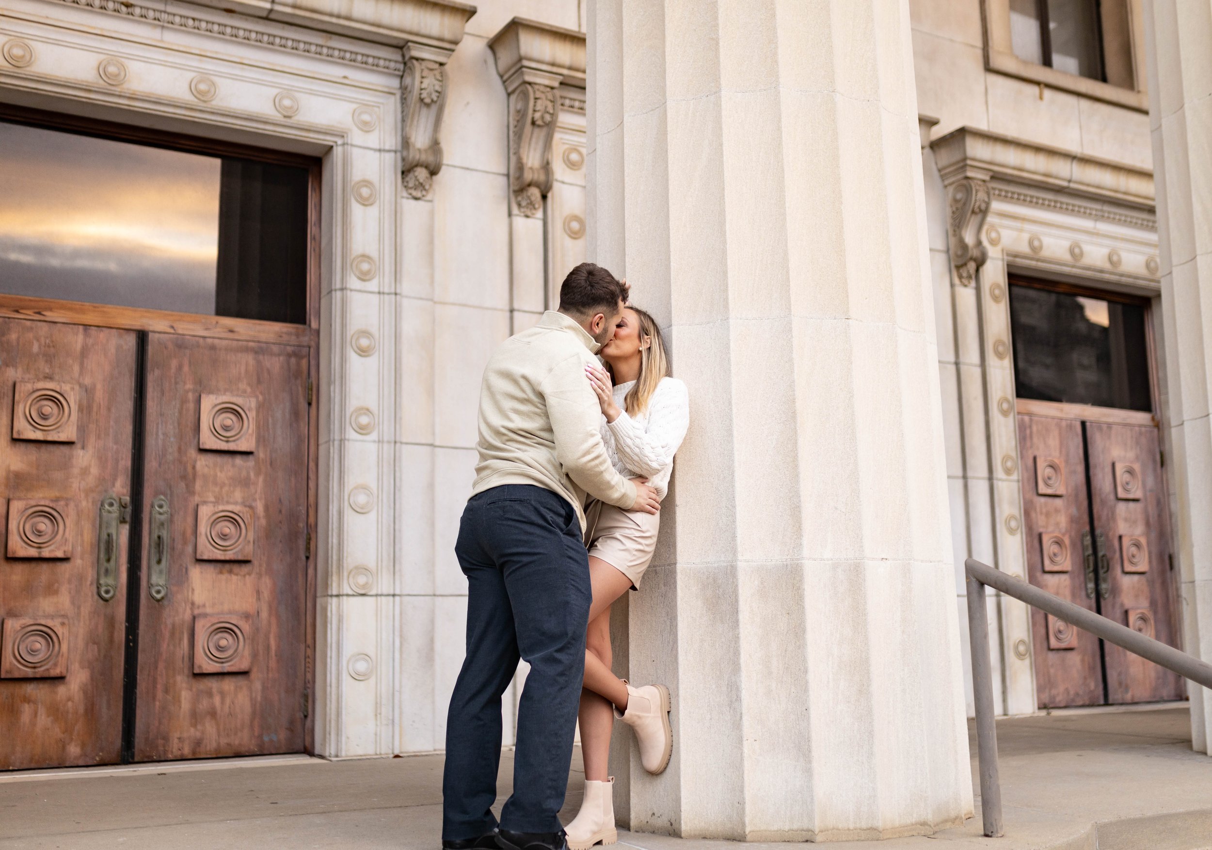 A couple kissing outside an ornate building with large wooden doors, the woman leaning against a column wearing shorts and boots, the man leaning in for a kiss facing her.