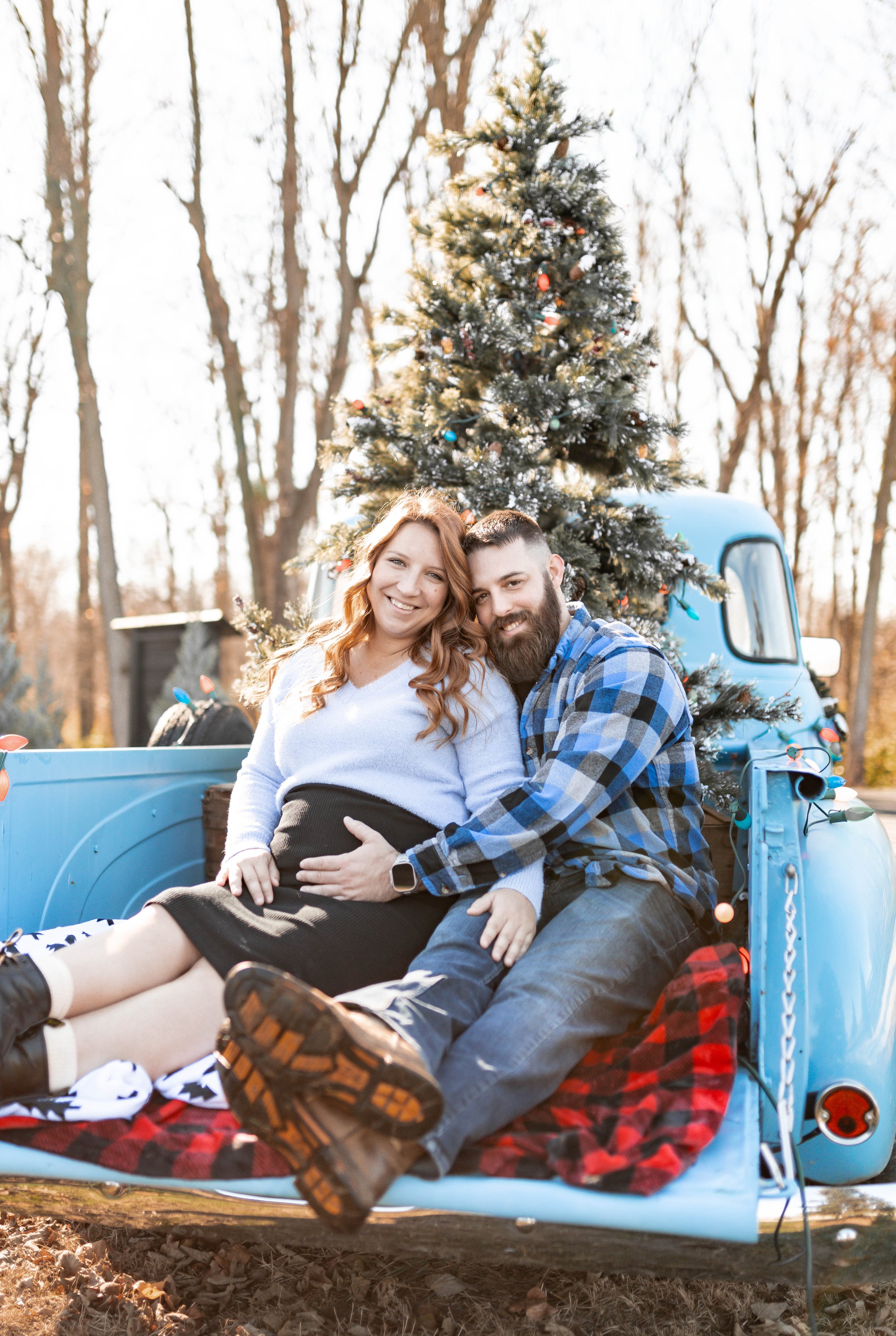 A smiling couple sitting on the back of a vintage blue truck with a Christmas tree, outdoors during daytime.