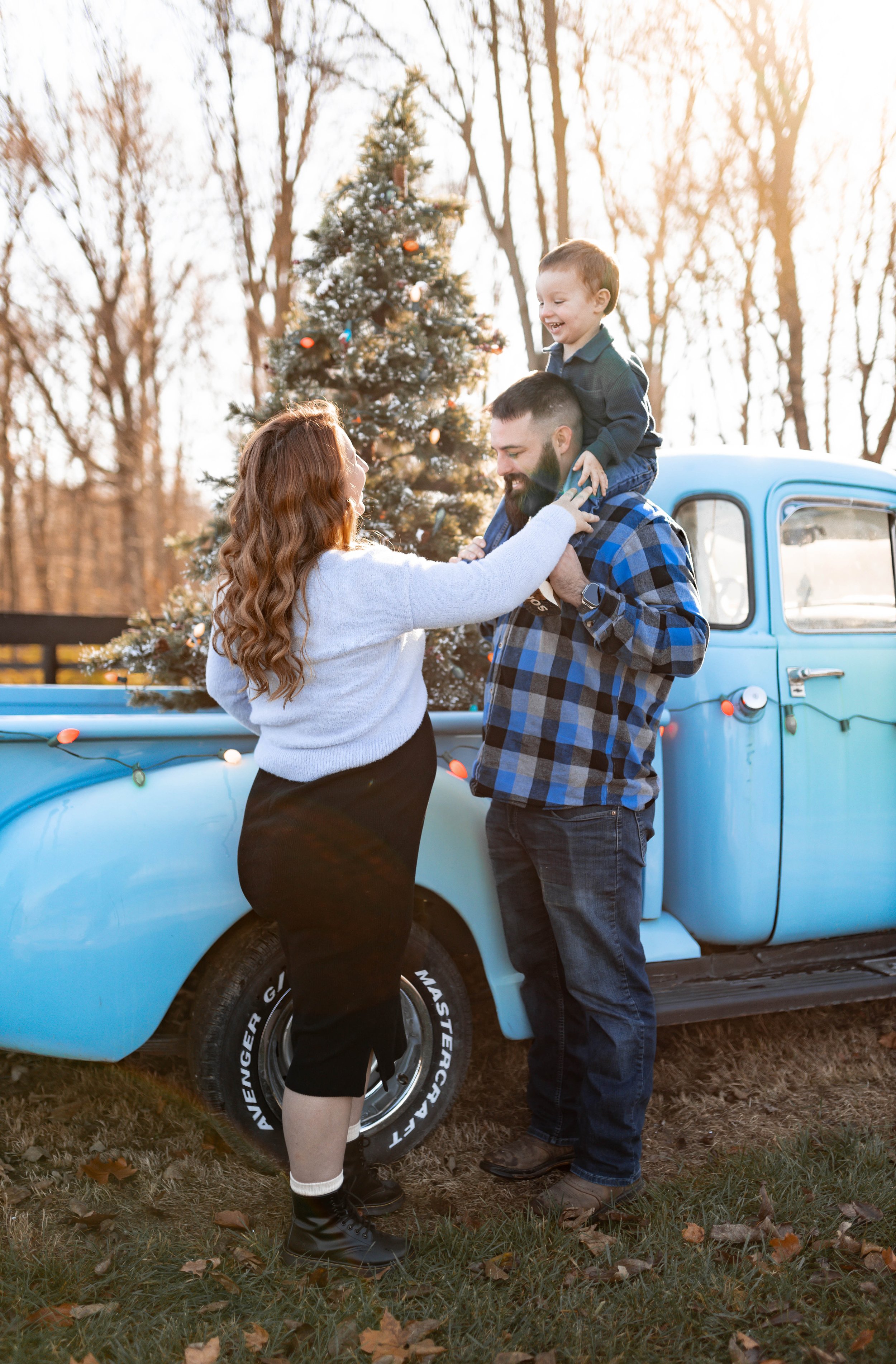 A family of three celebrating Christmas outdoors next to a decorated tree, with the father holding their young son on his shoulders, as the mother interacts with them.