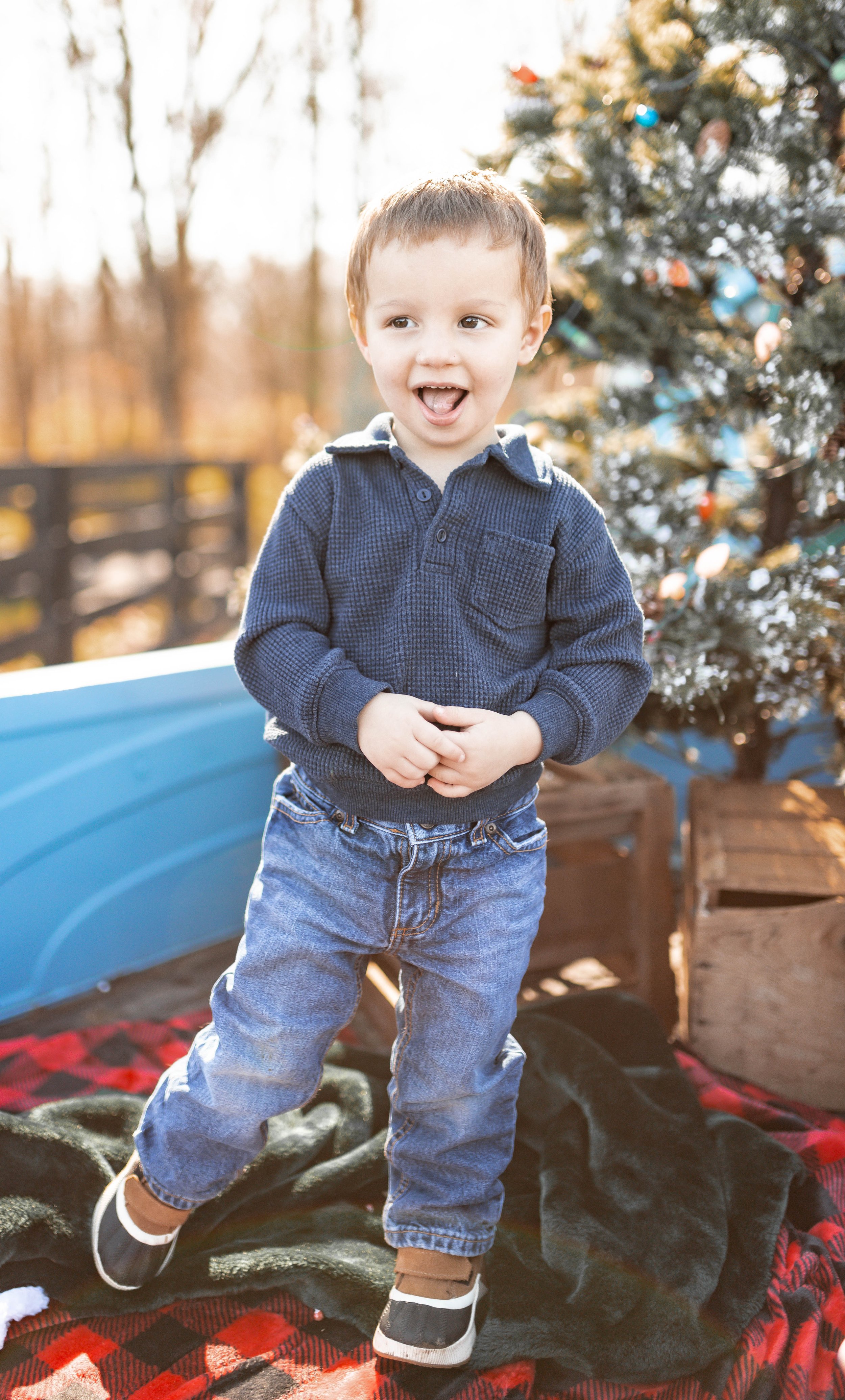 Smiling young boy in blue sweater and jeans standing outdoors near a decorated Christmas tree during daytime.