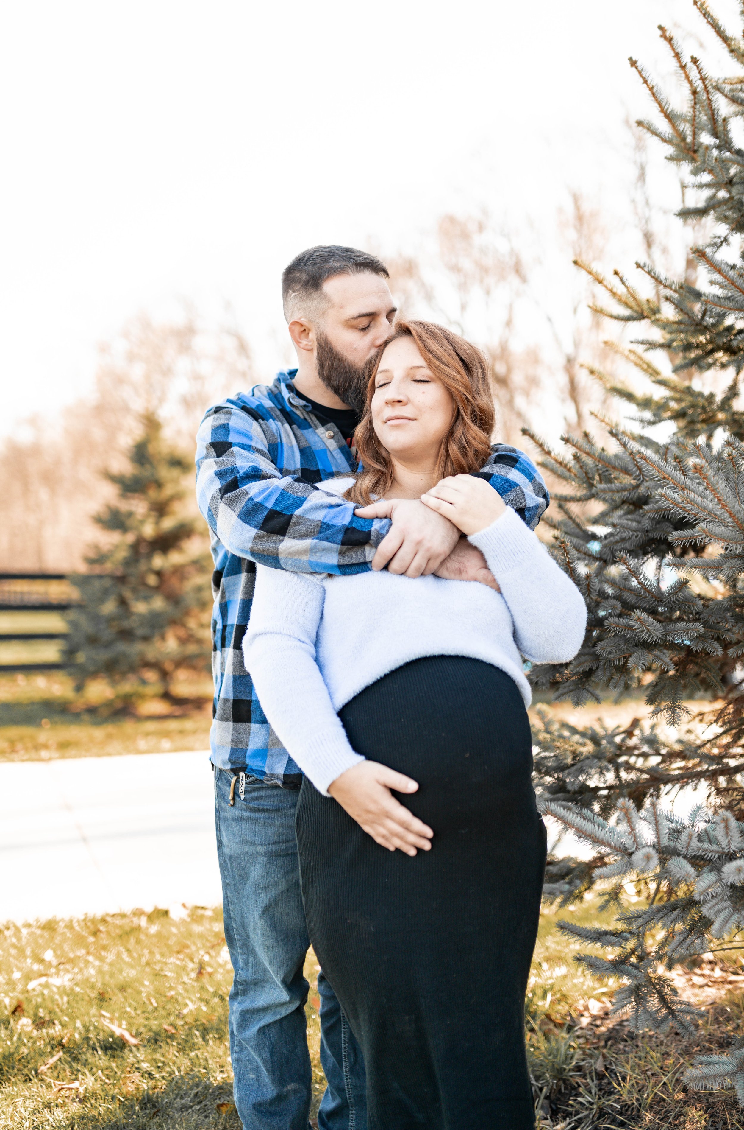 A man with a beard and a woman with red hair stand outdoors near a pine tree, embracing with the man kissing the woman's forehead. The woman is pregnant and wearing a black skirt and a white sweater, while the man wears a blue and black plaid shirt a