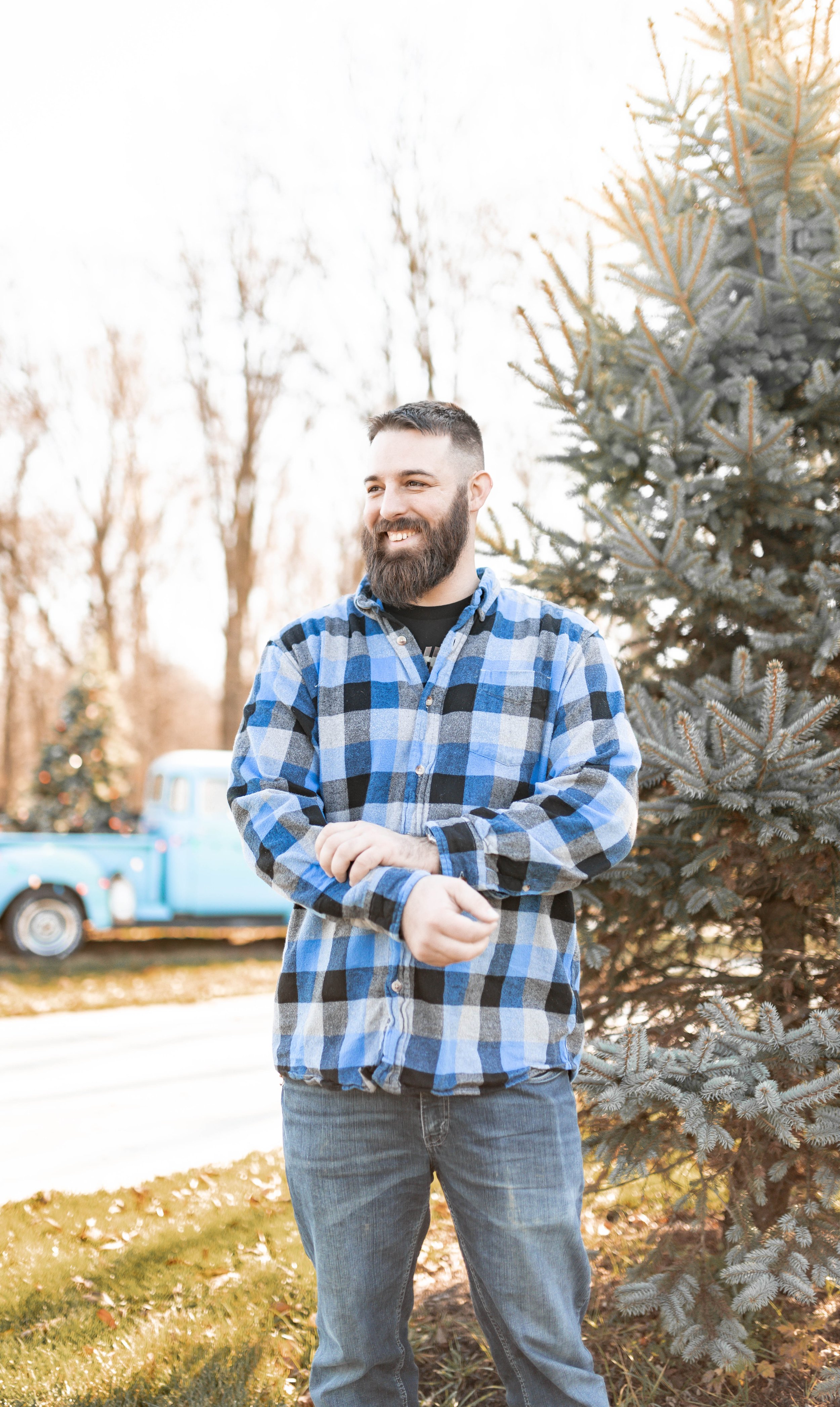 A man with a beard and short hair wearing a blue and black plaid shirt and gray jeans standing outdoors near a Christmas tree, smiling, with a vintage blue truck in the background.