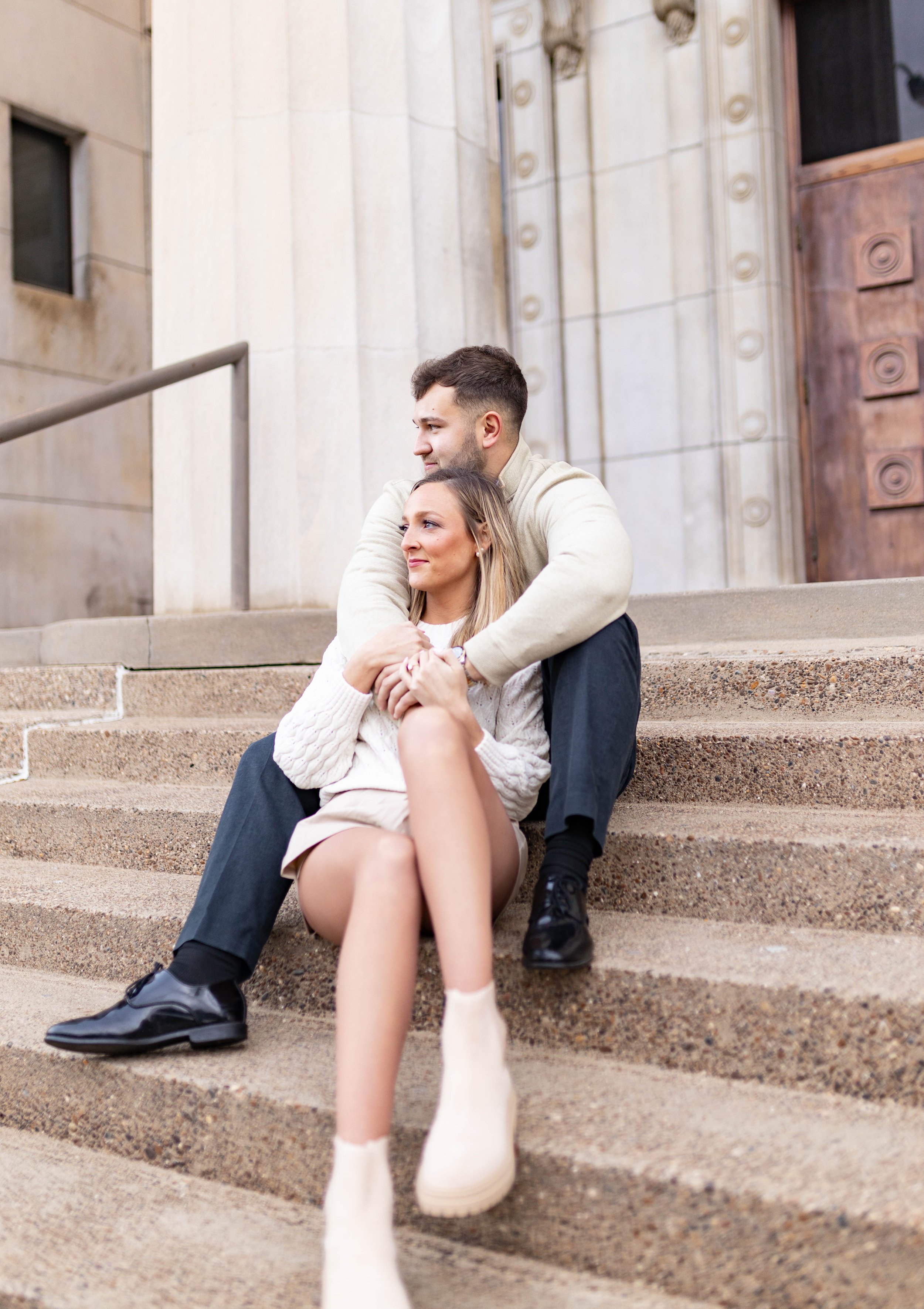 A couple sitting on stone steps in front of a building, embracing each other and looking off into the distance.