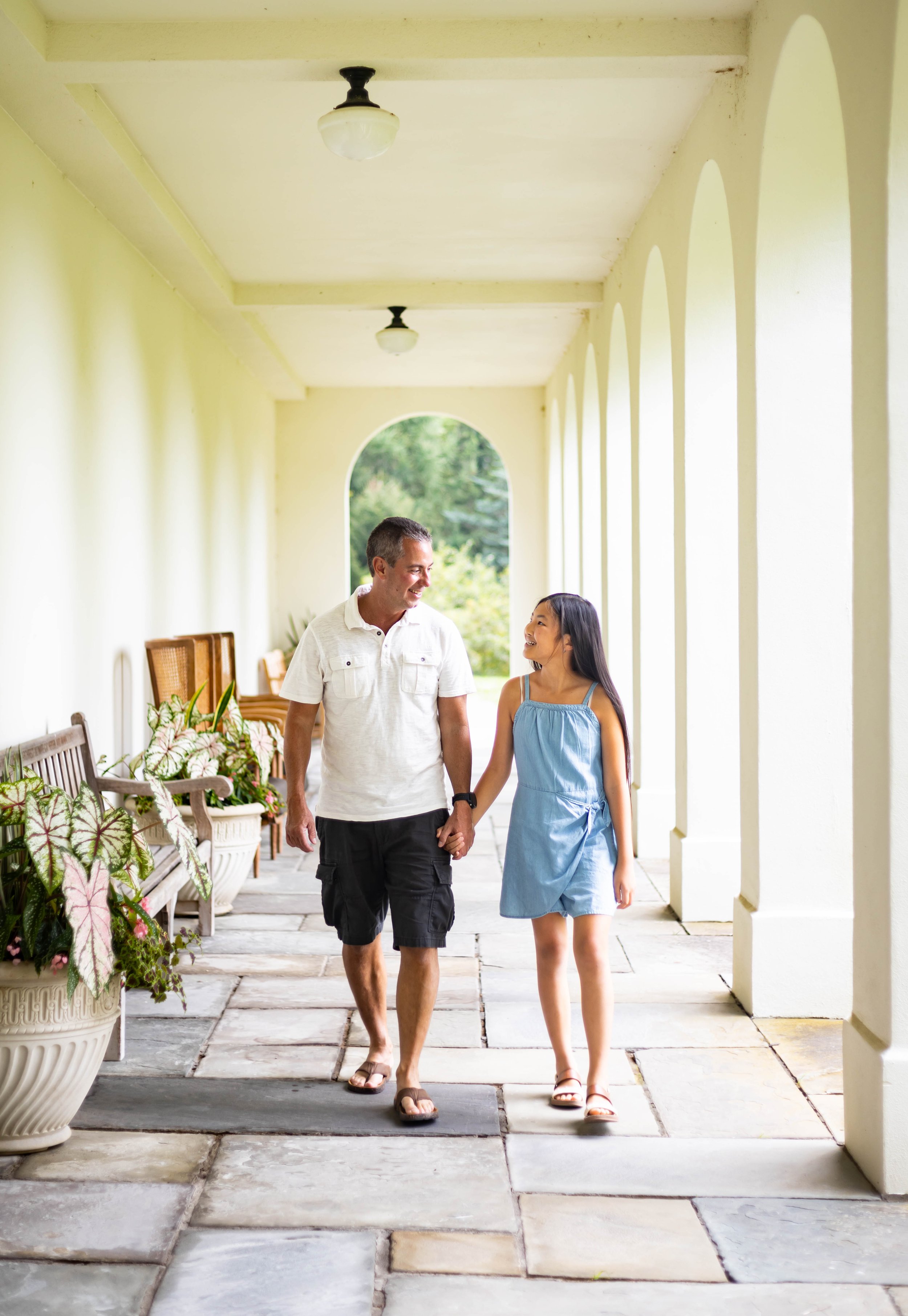 A man and a girl walking hand in hand down an arched outdoor corridor, smiling and looking at each other, with sunlight and plants in the background.