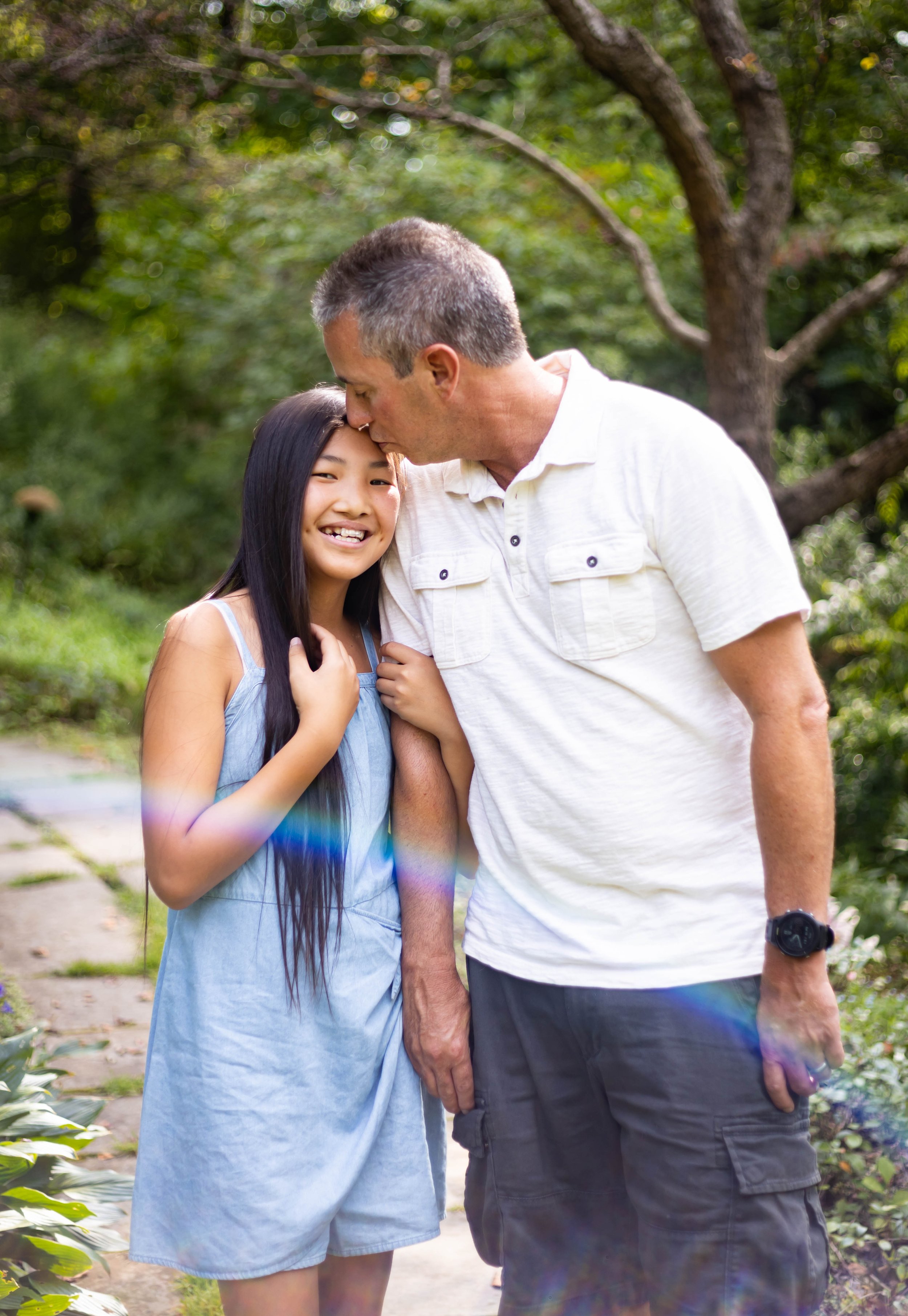 A man giving a kiss on the forehead to a young girl with long black hair in a garden with green trees and plants.