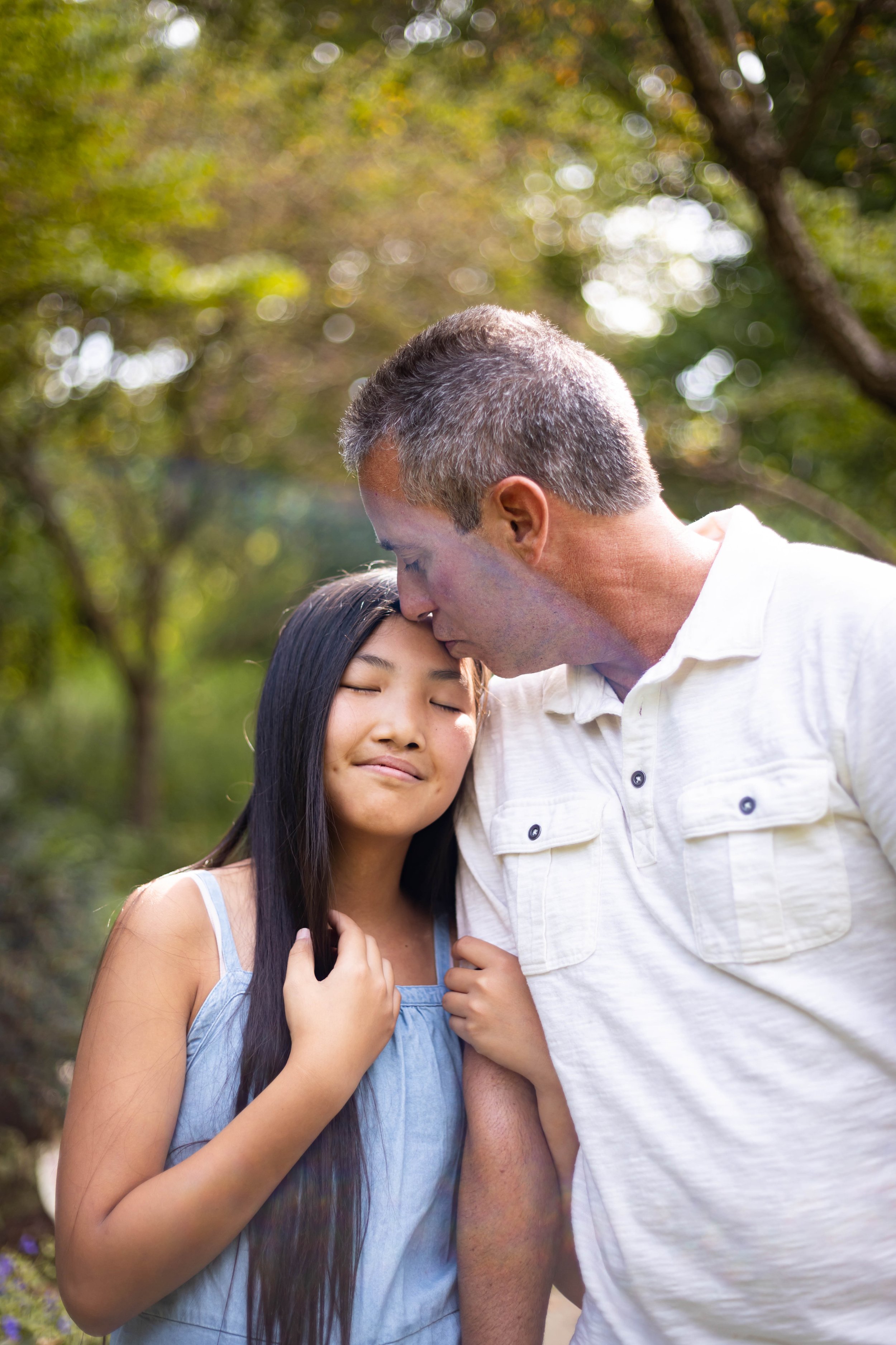 An older man kissing a young girl on the forehead outdoors with trees in the background.