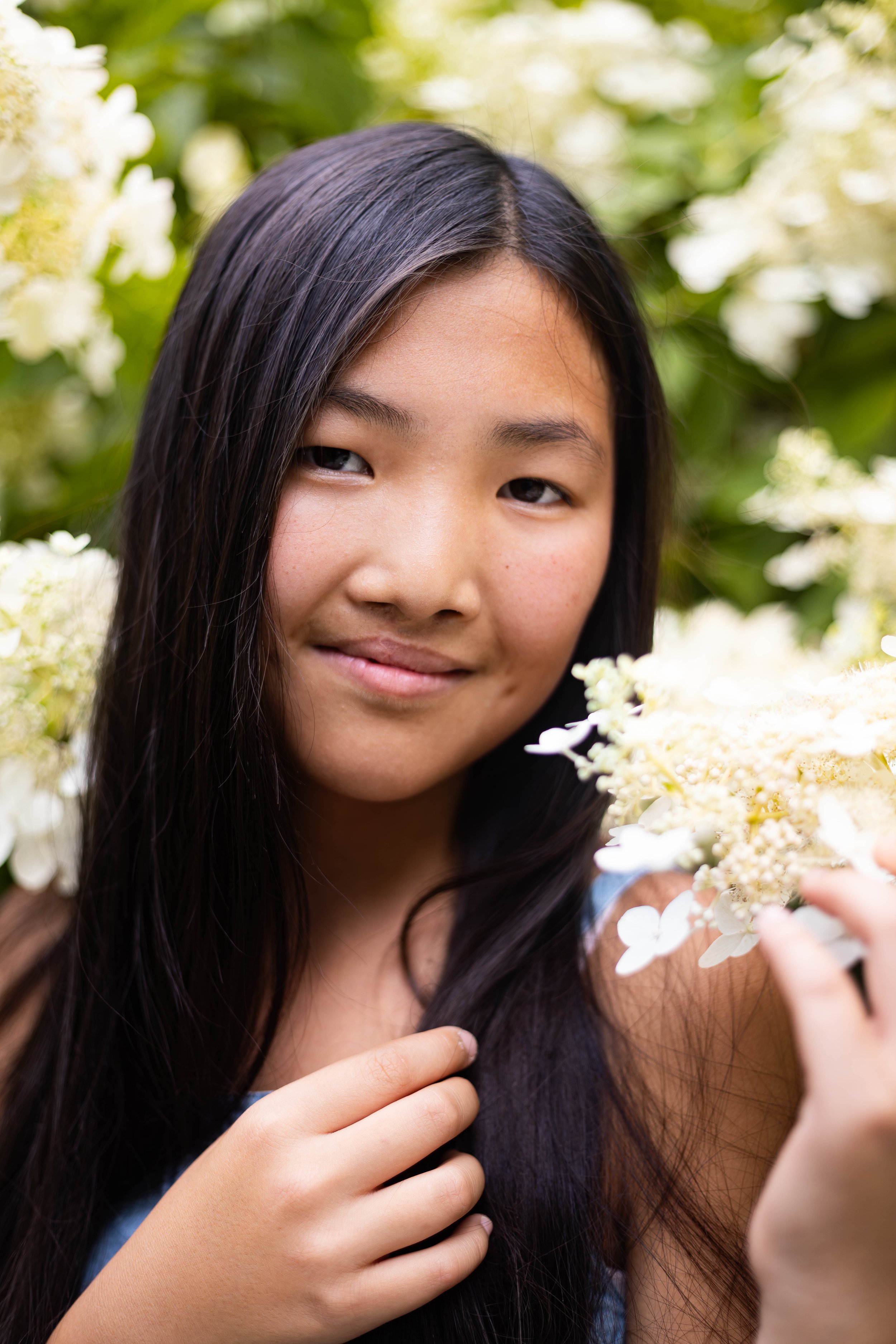 A young girl with long dark hair holding a white flower, standing among green leaves and white flowers.