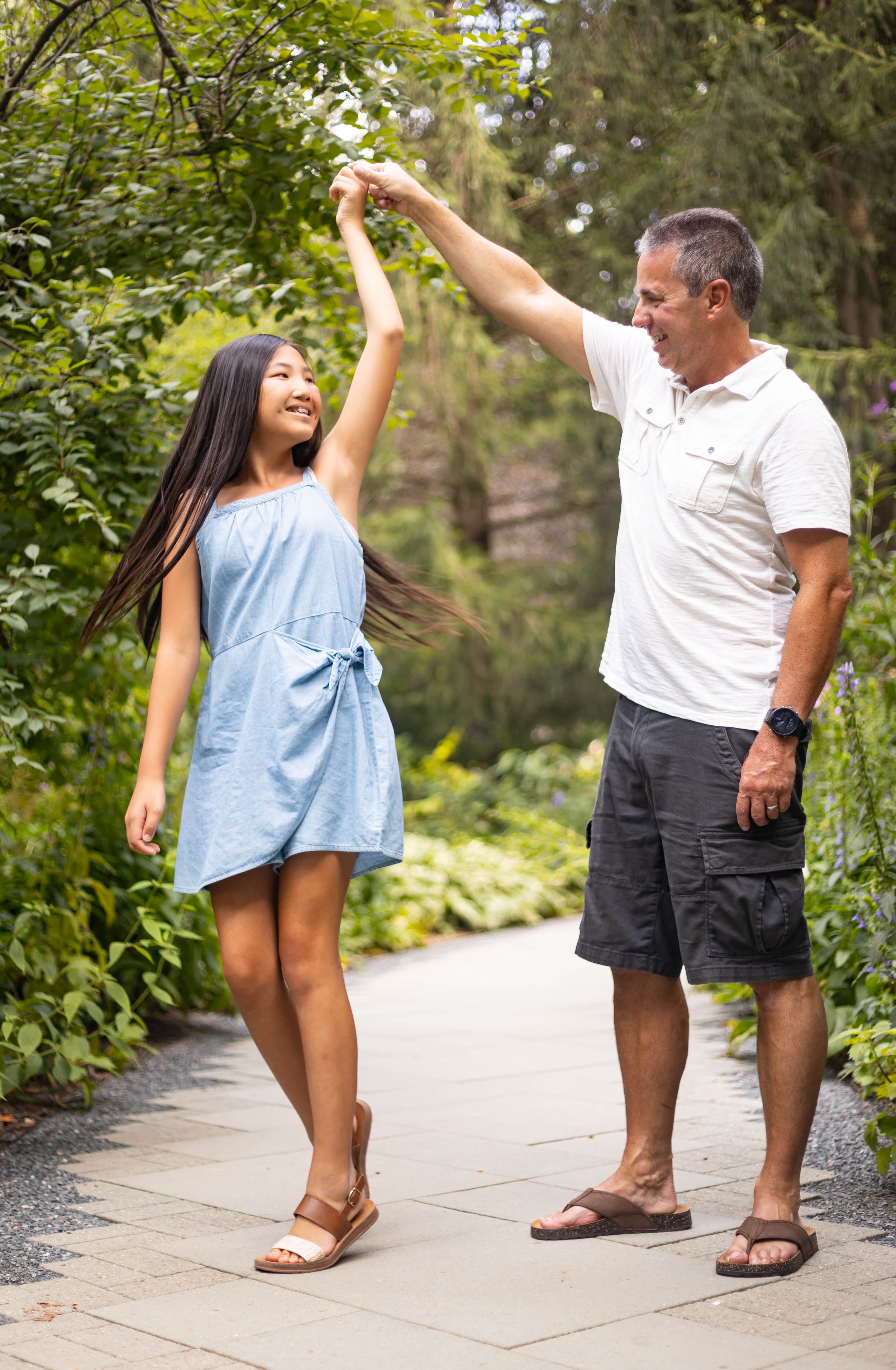 A girl and an older man dancing and smiling on a garden path surrounded by lush greenery.